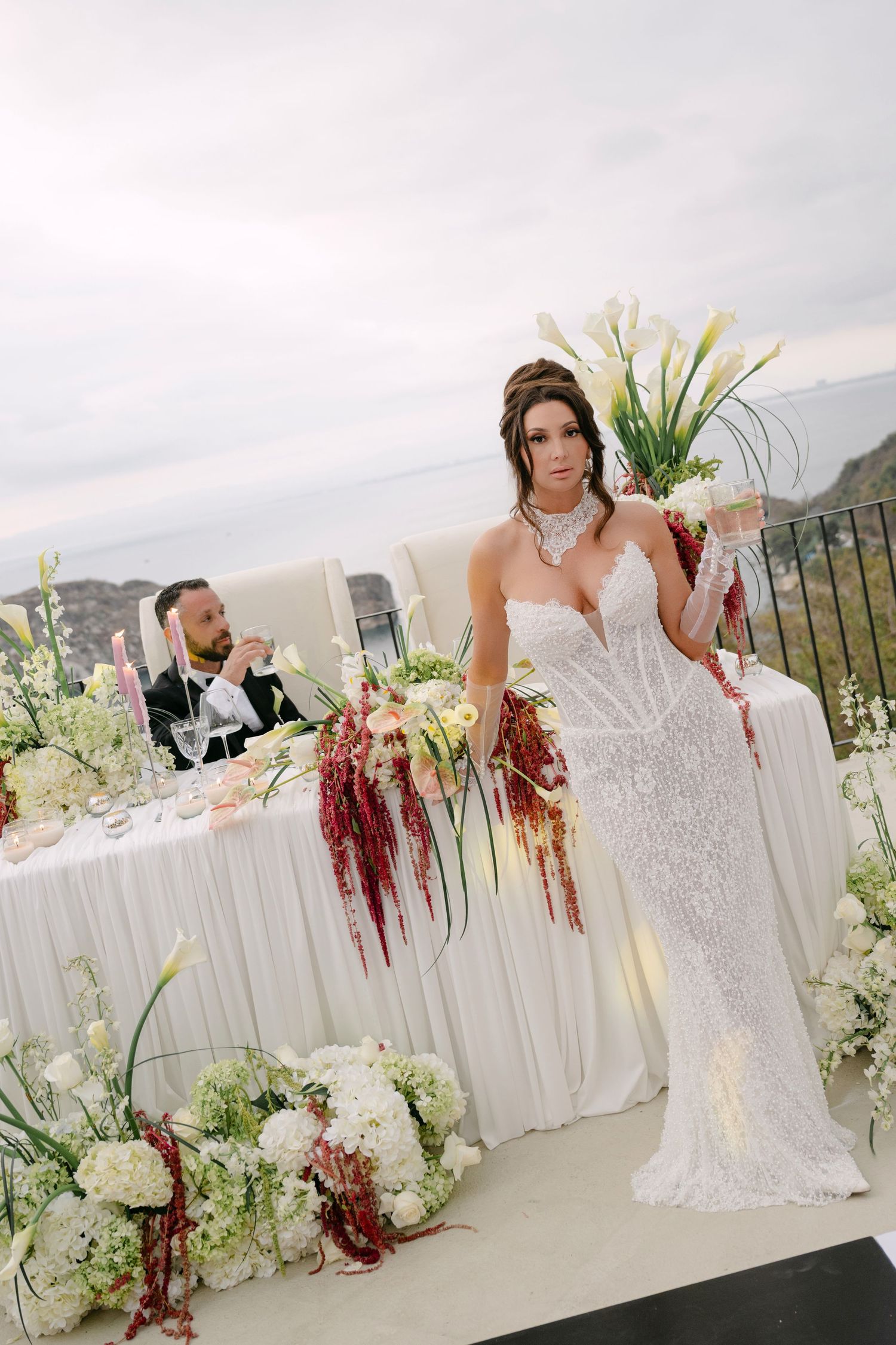 Elegant bride in a white sequin gown holding florals at a decorated outdoor wedding reception table with ocean views.