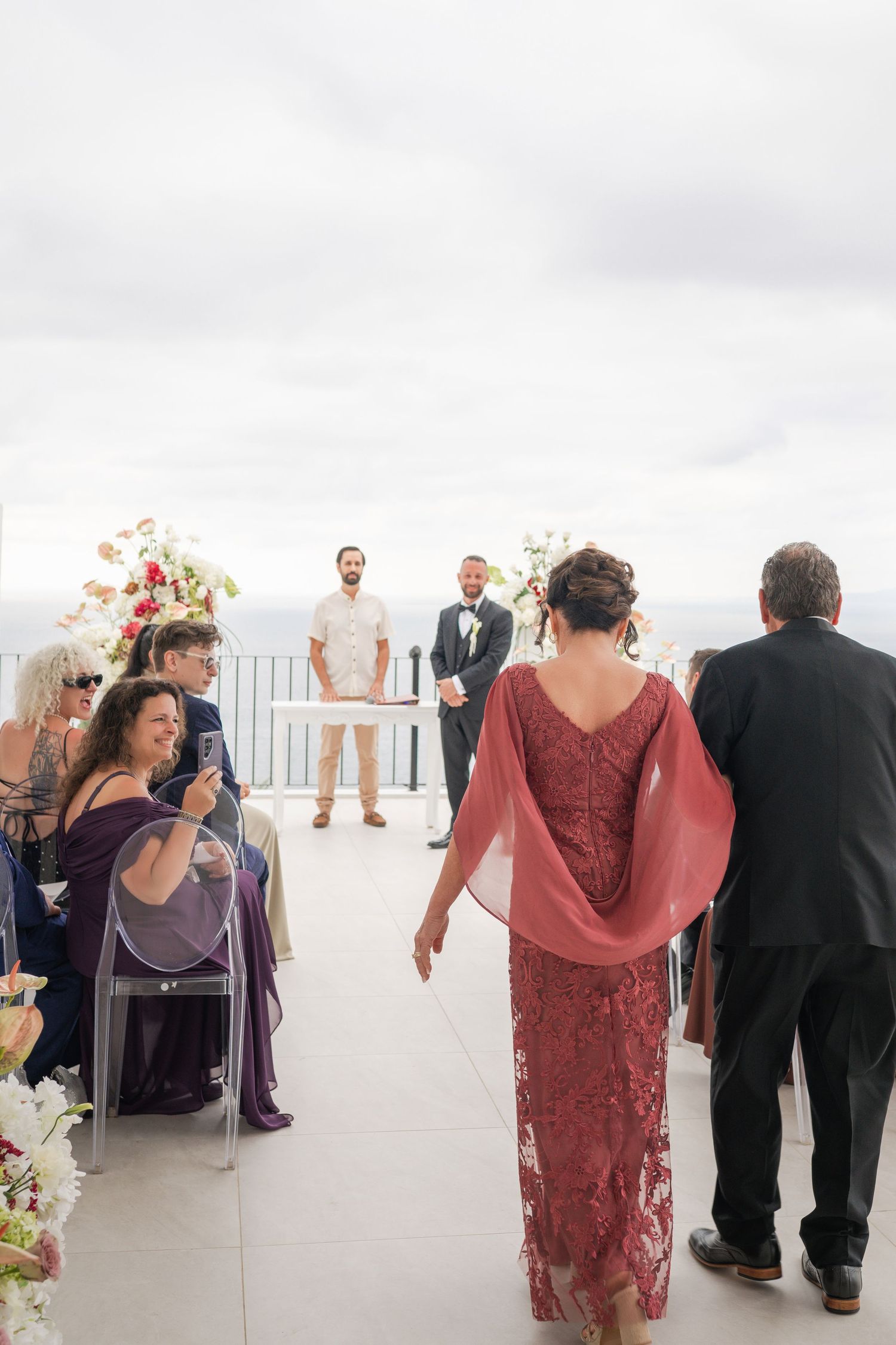 Bride walks down the aisle at an outdoor wedding ceremony on a terrace with ocean views.