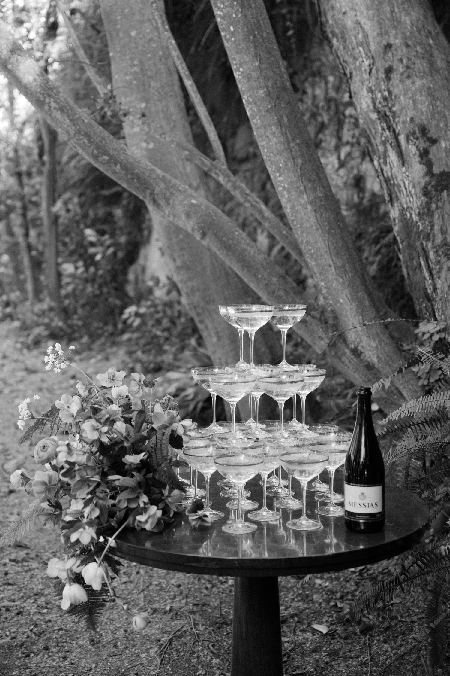 Black and white photo of elegant champagne tower on round table beside bottle and floral arrangement.