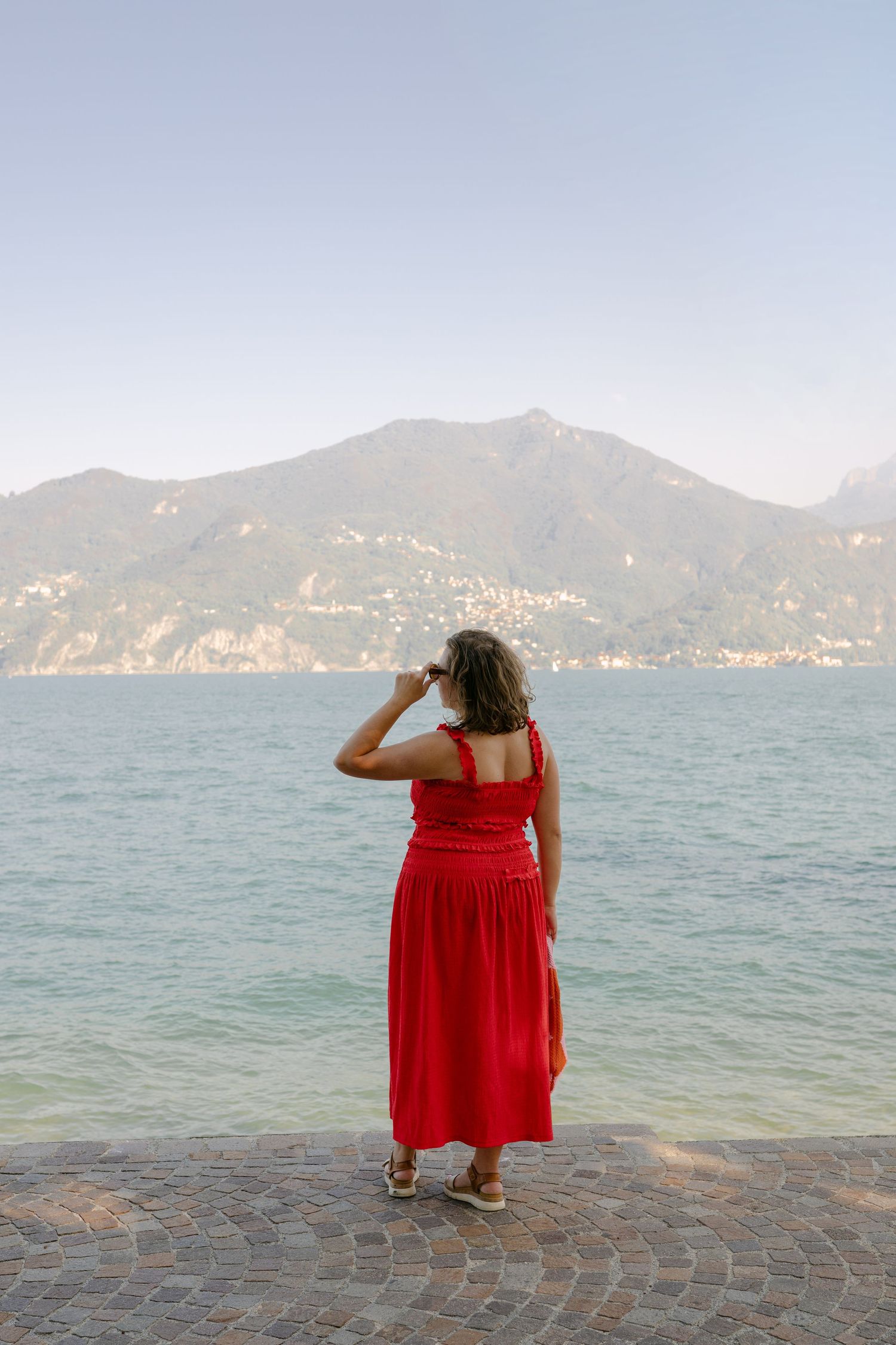 Woman in red dress standing on cobblestone pier gazing at turquoise lake with mountain backdrop.
