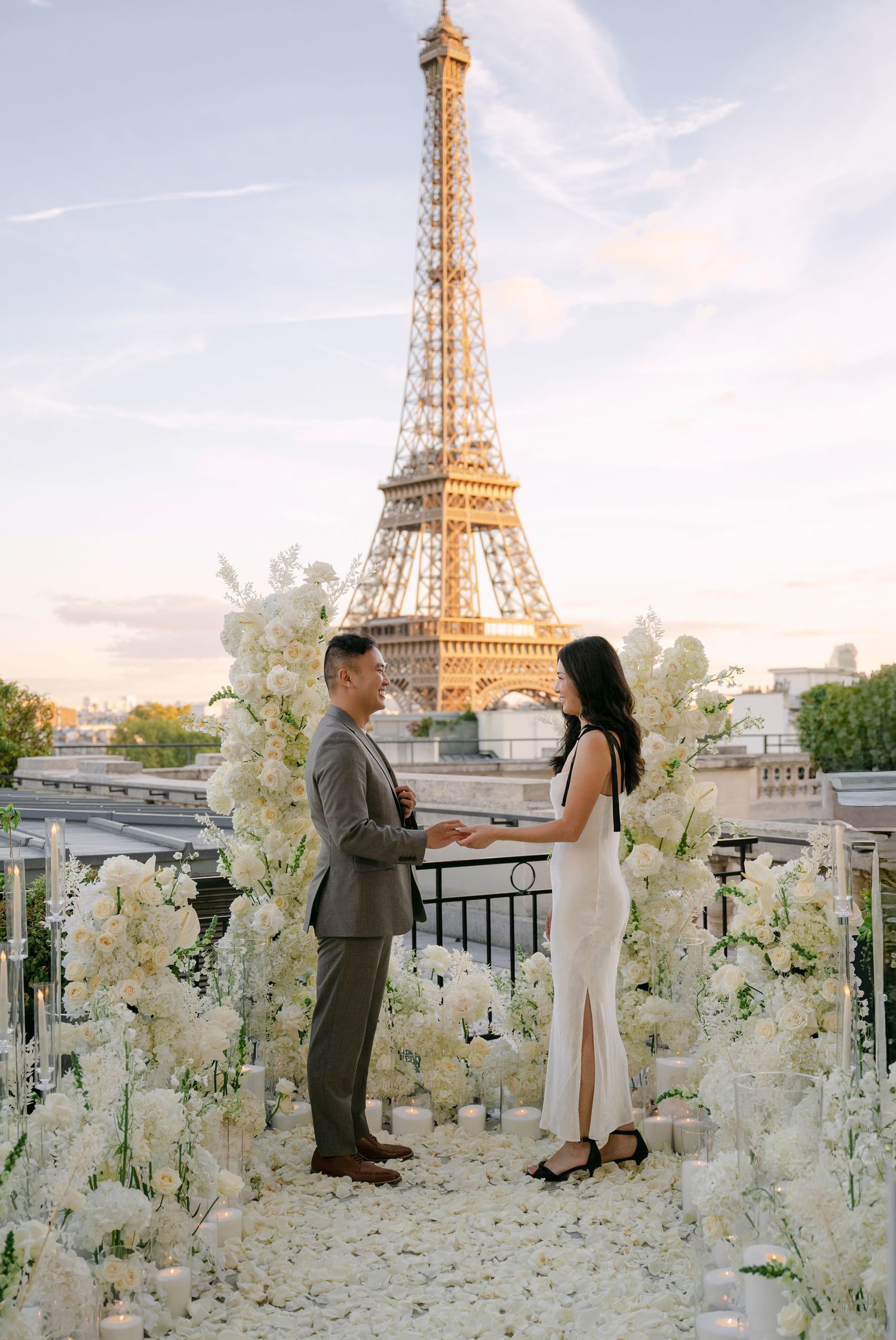 Couple holding hands during a romantic Paris rooftop proposal surrounded by white flowers with the Eiffel Tower glowing at sunset.