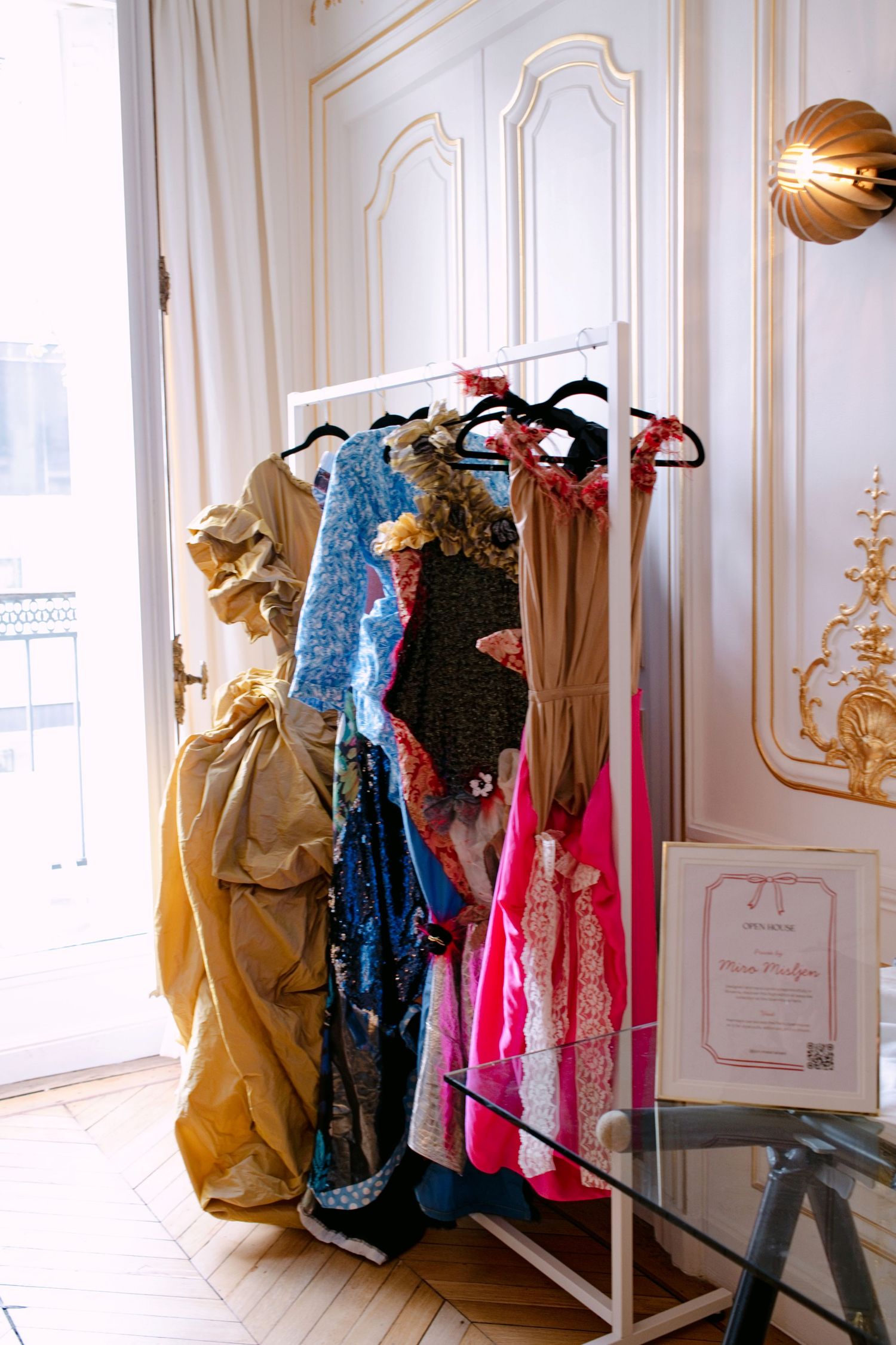 Colorful designer dresses displayed on a clothing rack in a bright Parisian apartment with gold trim.