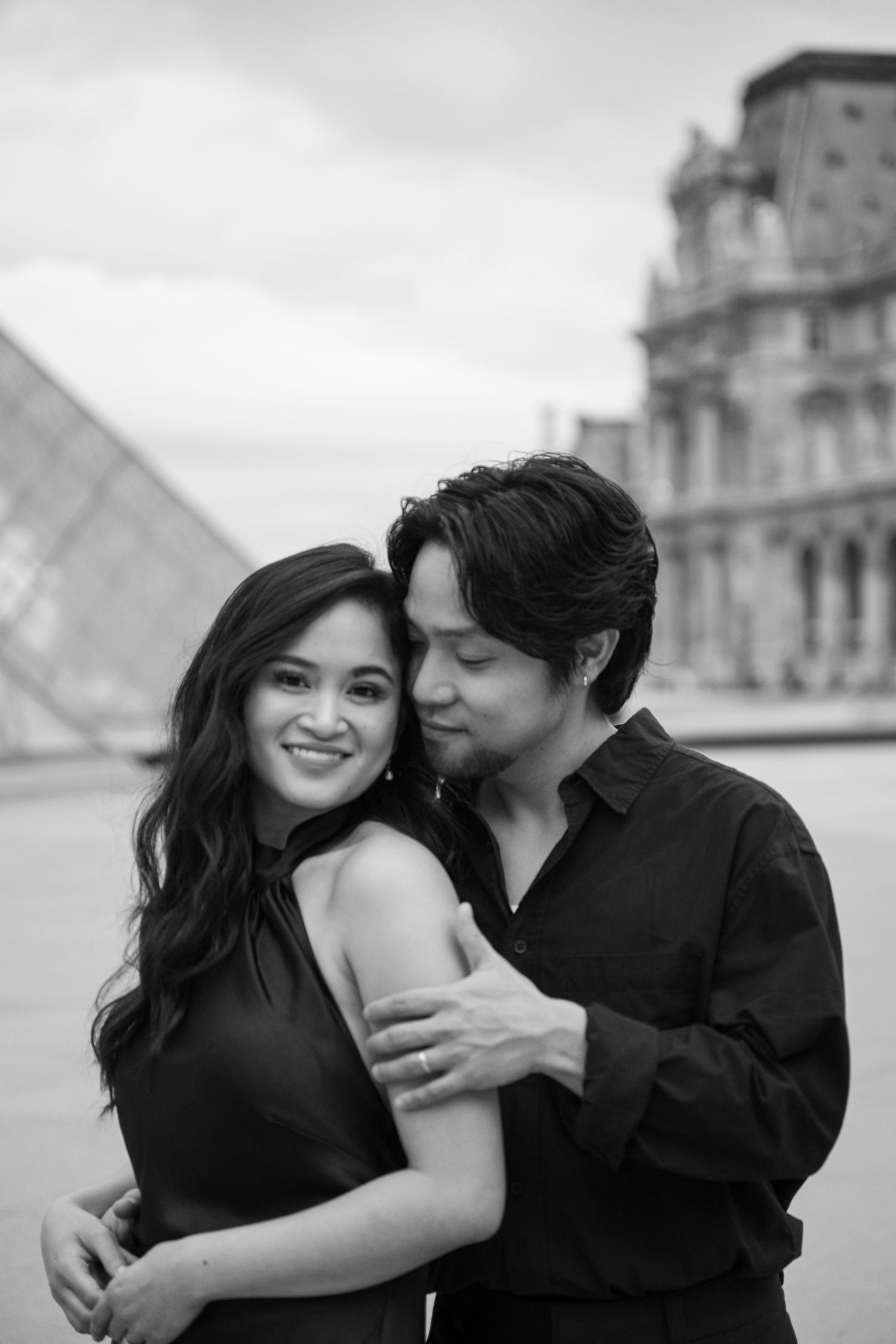 Black and white photo of a couple embracing in front of the Louvre pyramid in Paris.
