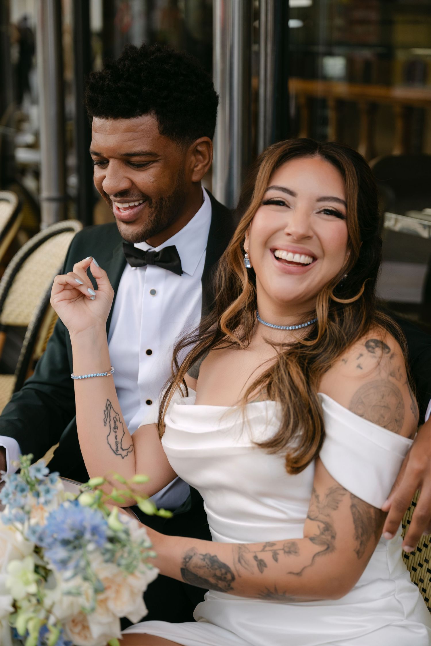 Laughing tattooed bride in off-shoulder gown sits with groom in tuxedo at Parisian cafe reception.