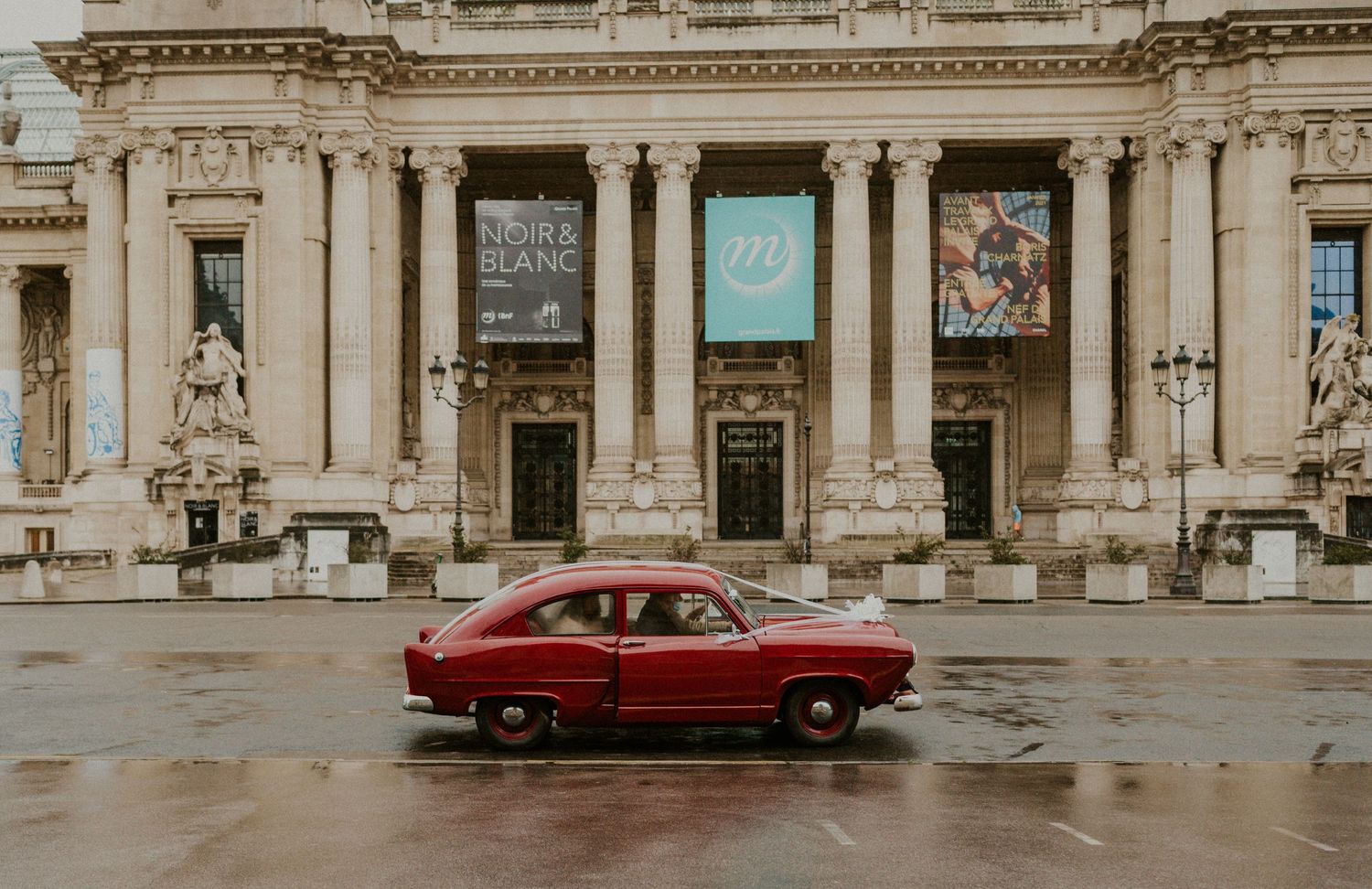 A vintage red car parked in front of a grand classical building with columns.