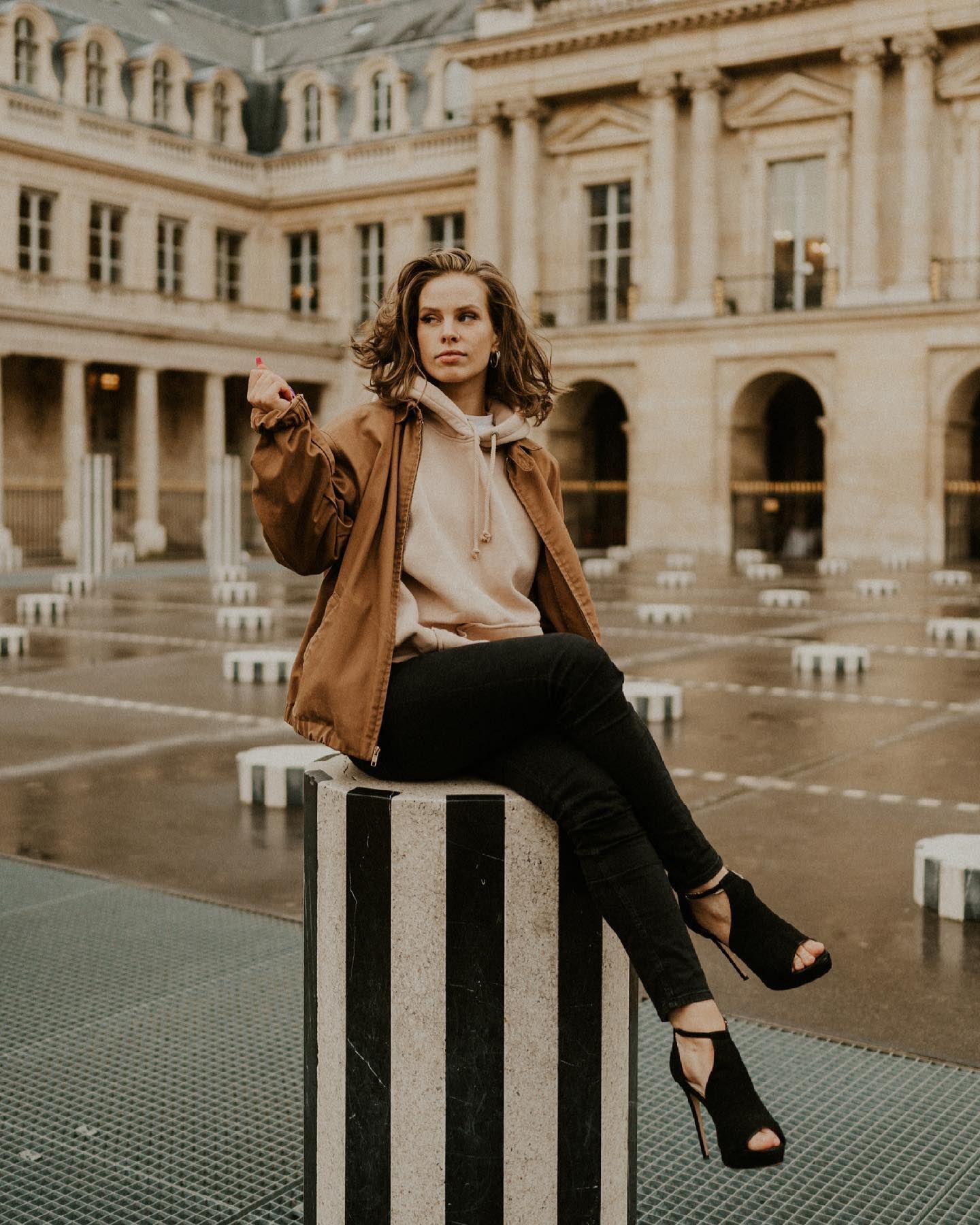 A person in casual chic attire poses on a striped column in a classical Parisian courtyard.