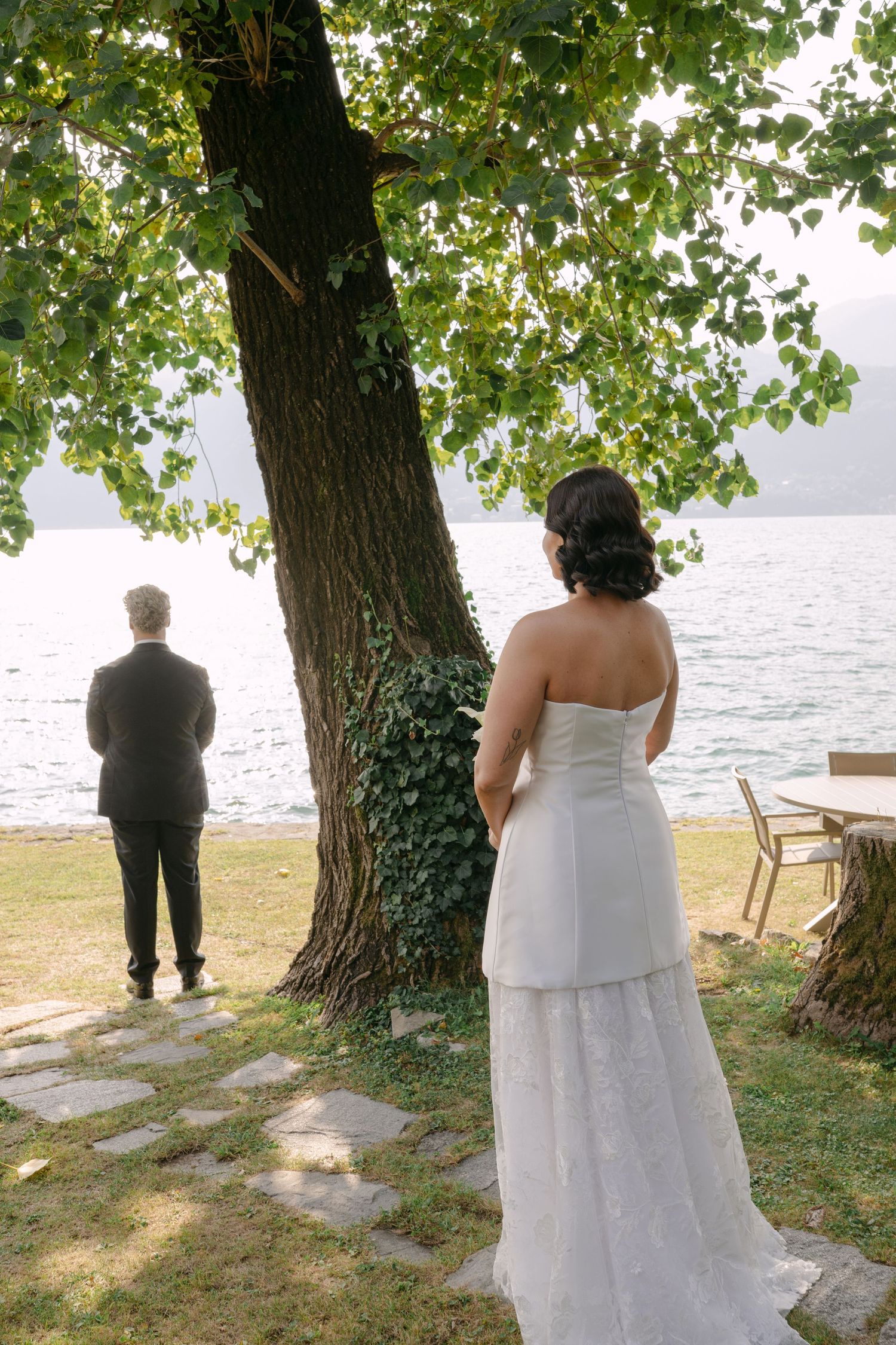 Bride in white strapless gown approaches groom near lakeside tree during outdoor wedding ceremony.