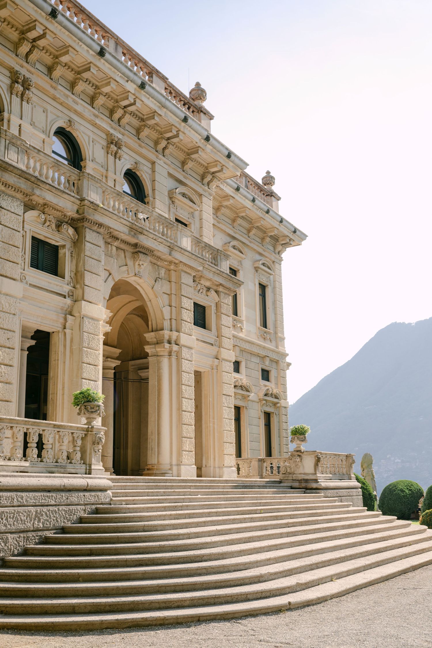 Grand Italian villa with ornate stone facade, arched entrance, wide staircase, and mountain backdrop under soft sunlight.