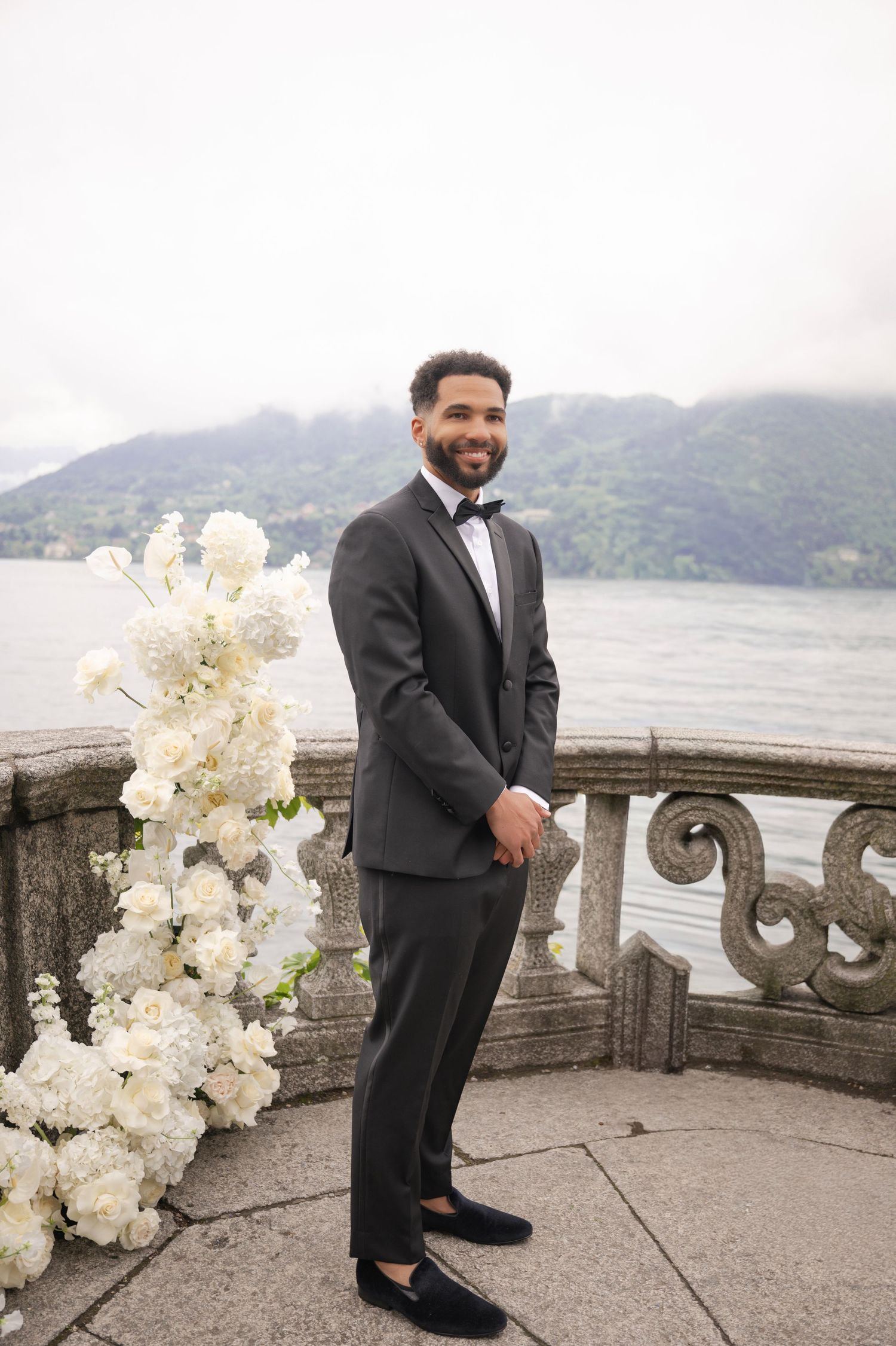 Groom in black tuxedo with bow tie stands by stone balustrade with white floral arrangement and lake backdrop.