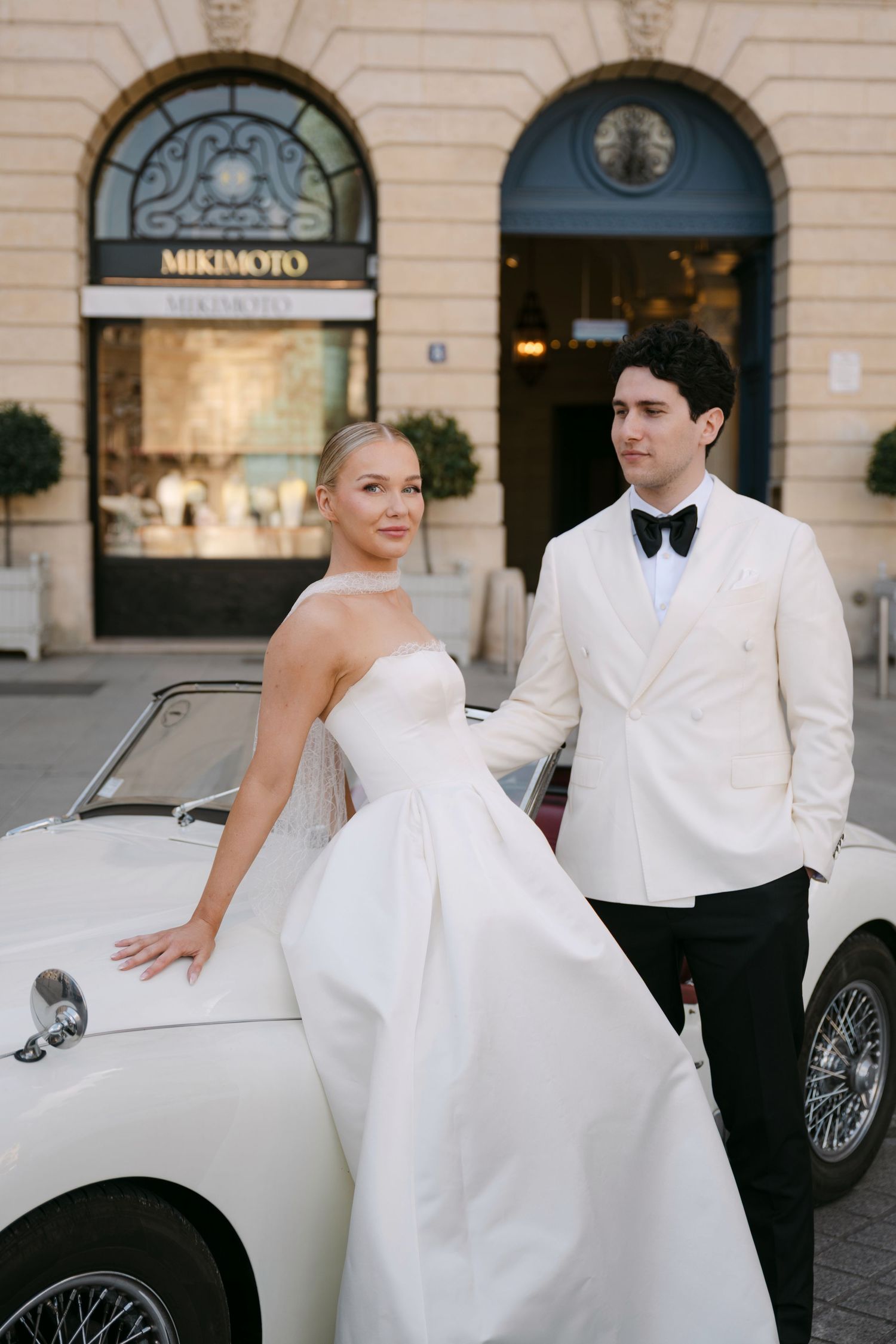 Elegant bride in strapless gown and groom in white tuxedo pose beside vintage white convertible outside Mikimoto.