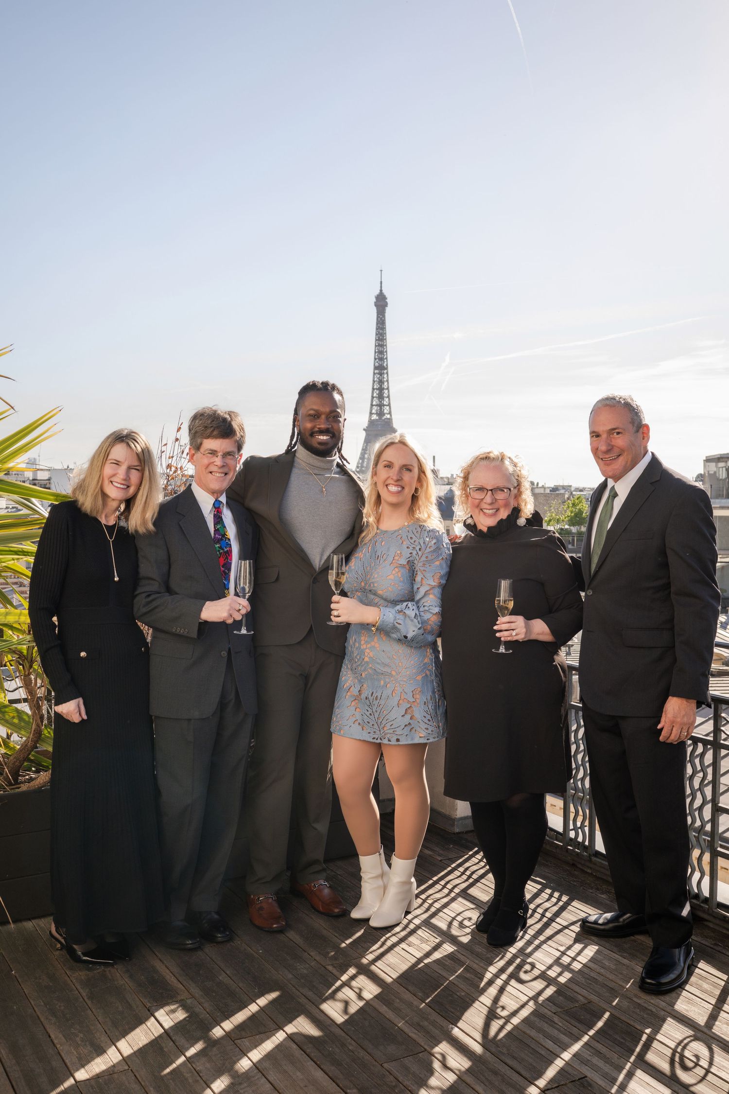 Six people posing with champagne glasses on Paris rooftop terrace with Eiffel Tower behind them.