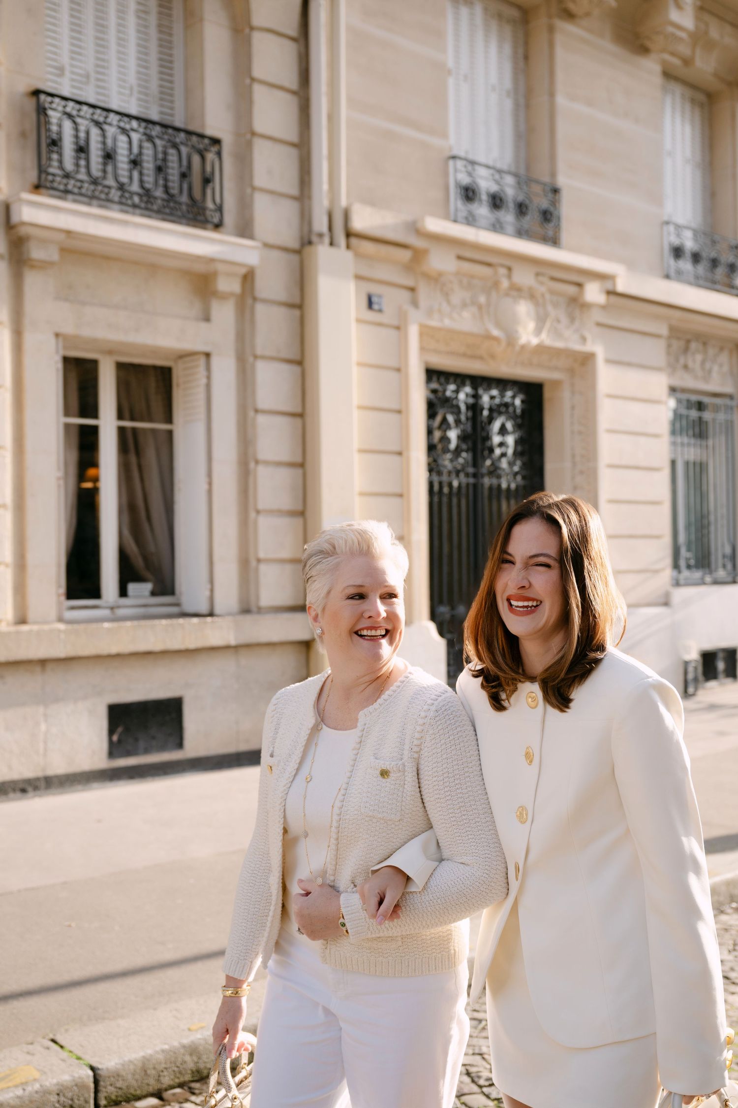 Two women in matching cream blazers laugh while walking along sunny Parisian cobblestone street.