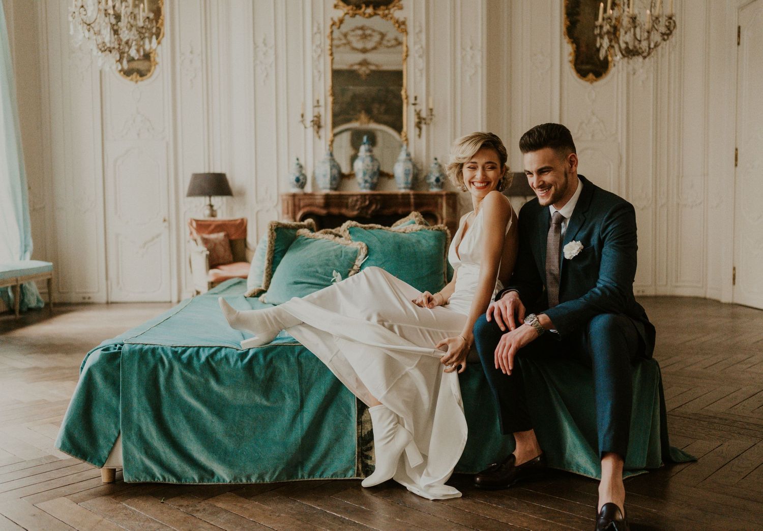 Elegant wedding couple resting on teal bedding in ornate white room with chandelier.