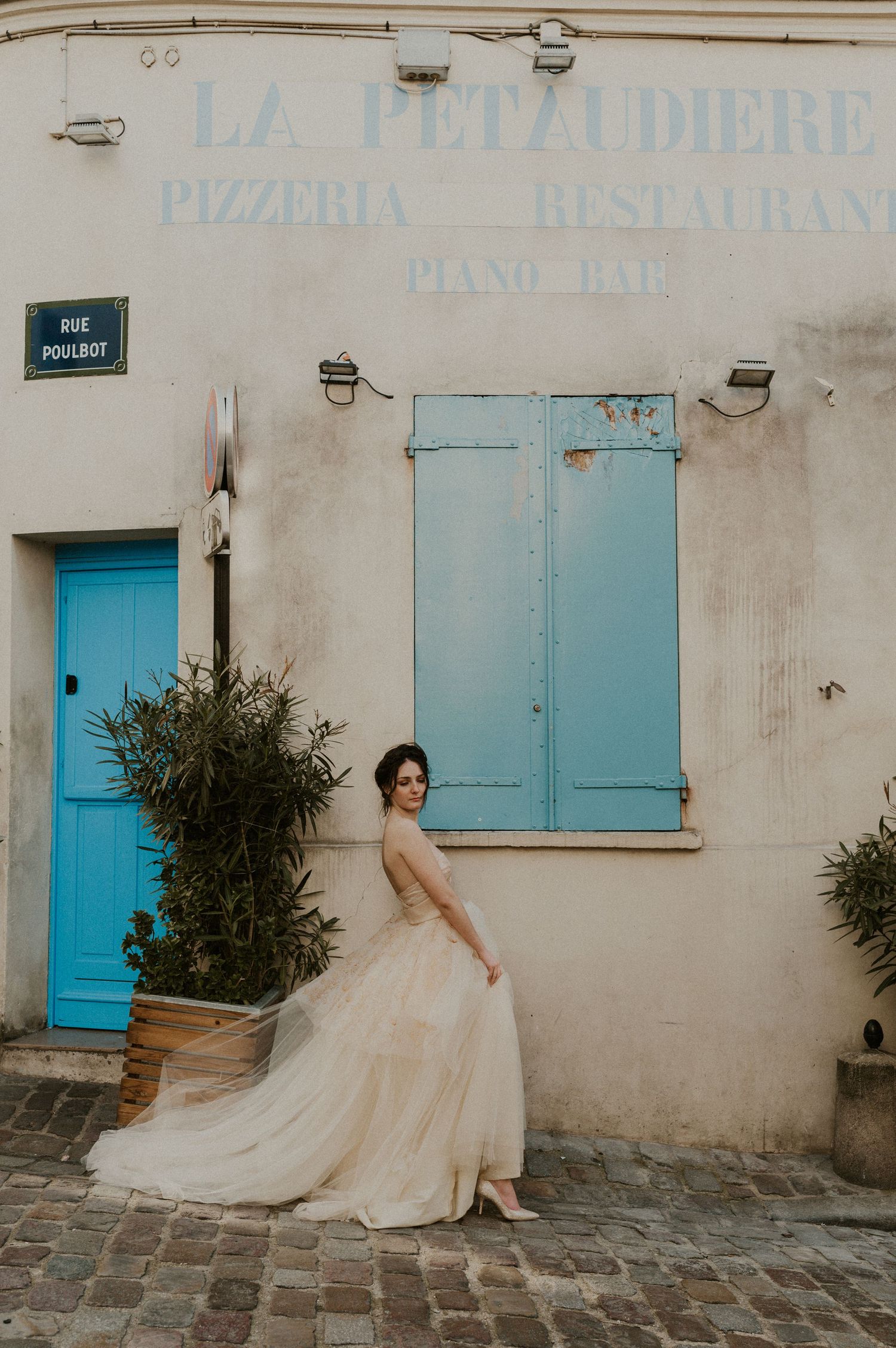 A bride in a flowing tulle gown poses against La Petaudiere restaurant with turquoise shutters.