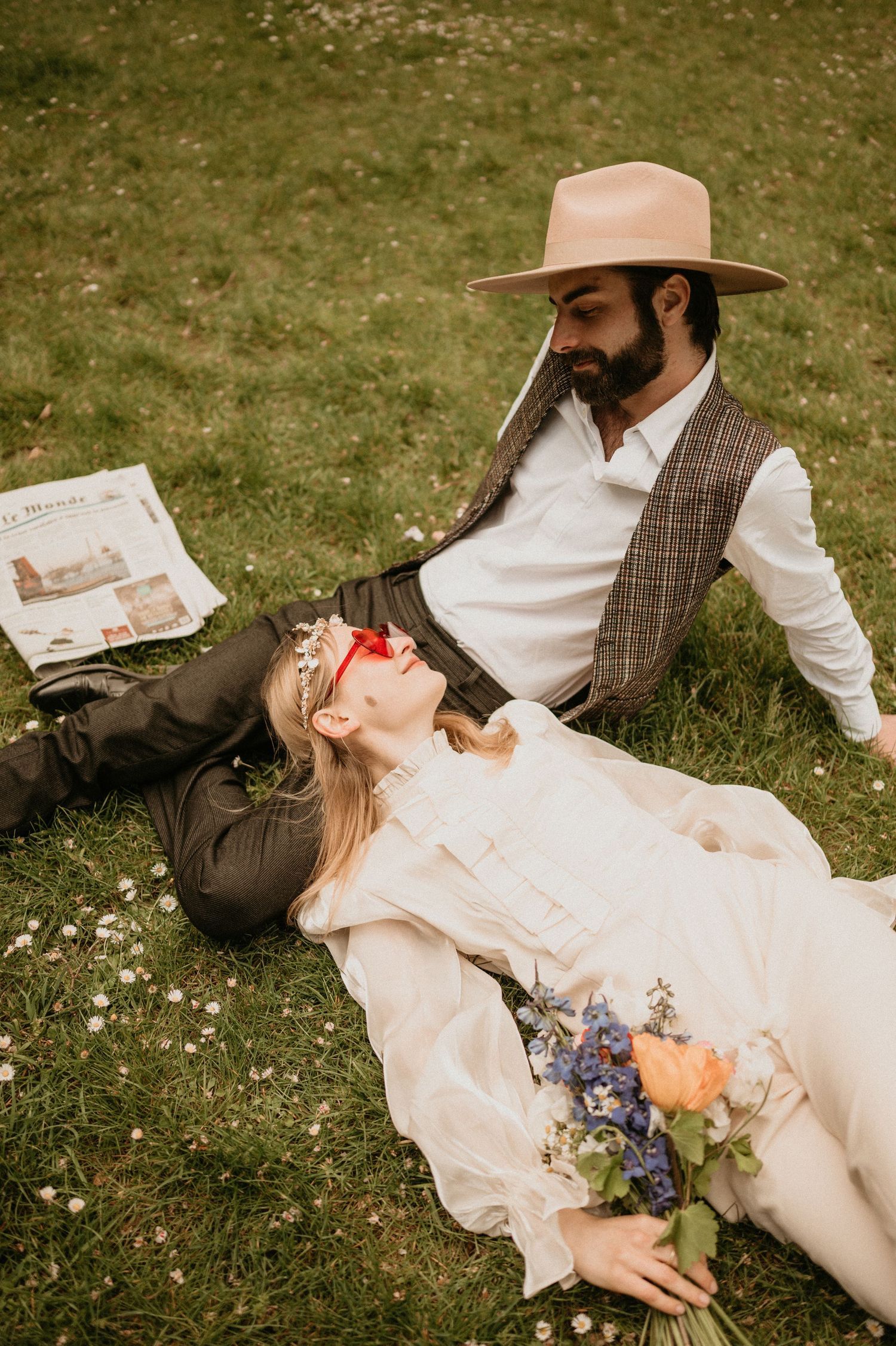 Couple in vintage clothing relaxing on grass with newspaper and colorful bouquet.