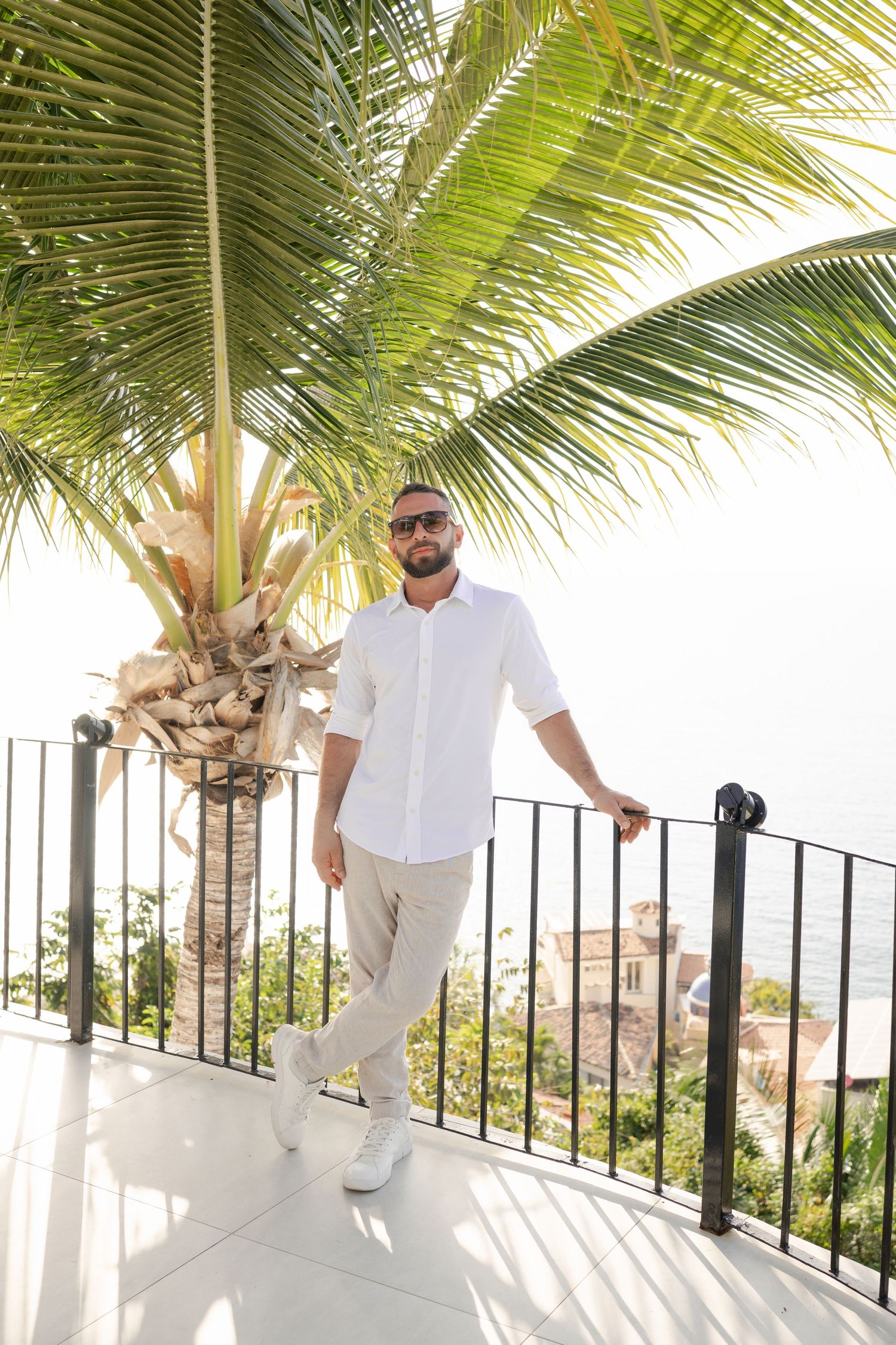 Man in white shirt and beige pants posing on a sunny terrace with palm tree and ocean view.