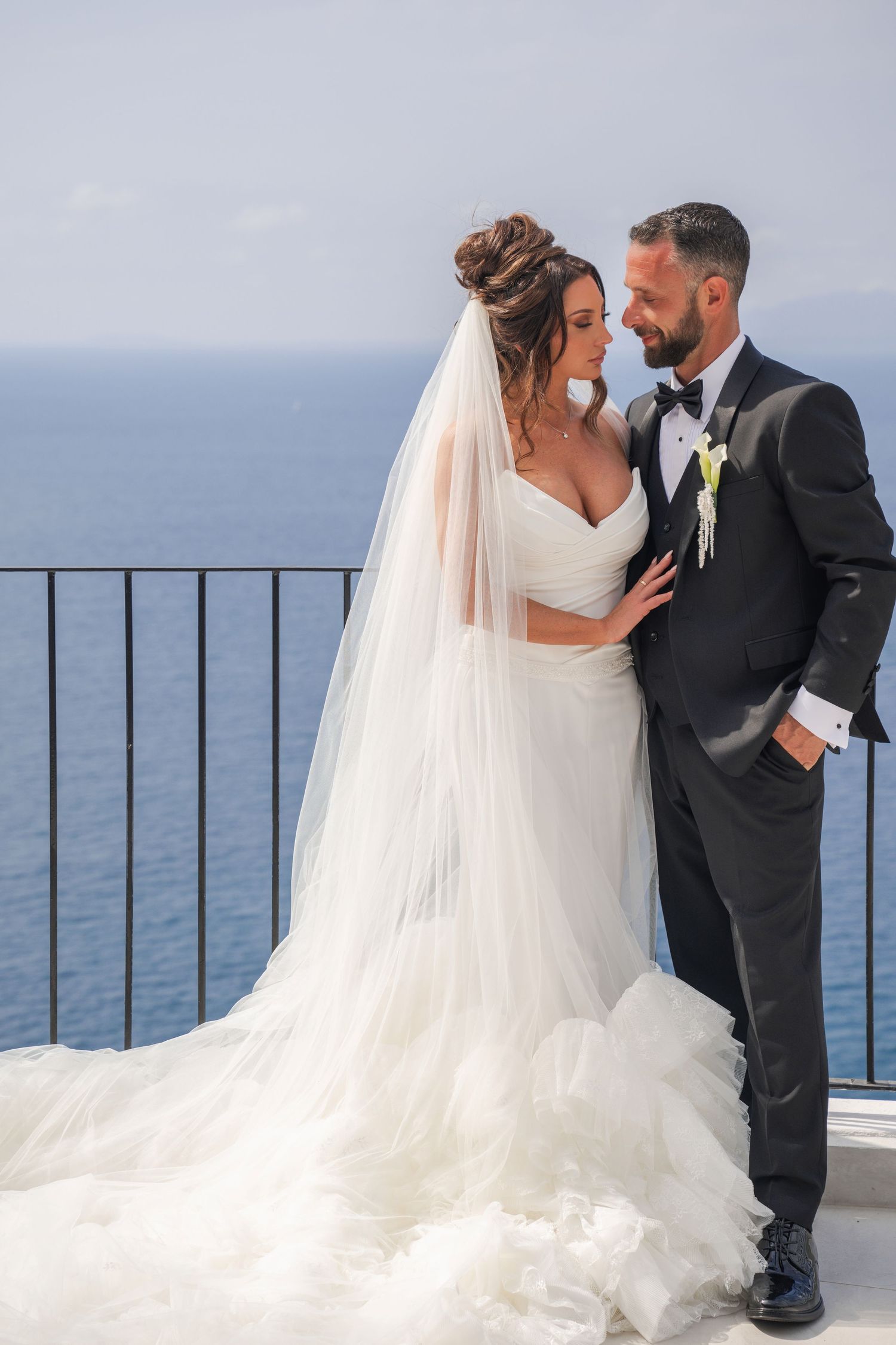 Bride and groom share a romantic moment on a seaside balcony, groom in black tuxedo.
