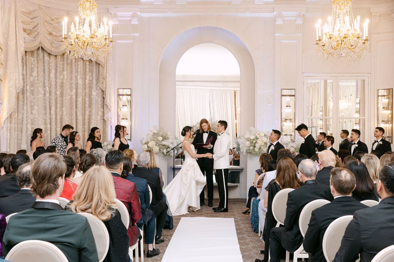 Wedding ceremony in elegant ballroom with bride and groom at altar, guests seated, crystal chandeliers overhead.