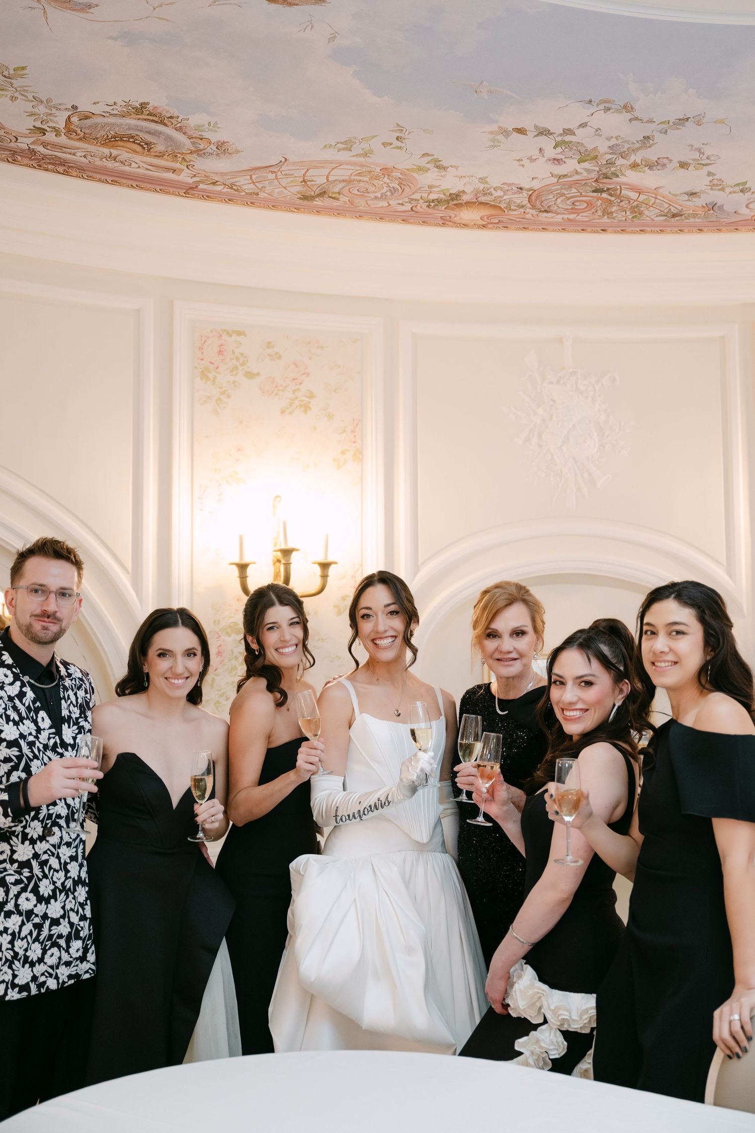 Bride in white gown poses with wedding party in black attire holding champagne in elegant ballroom.