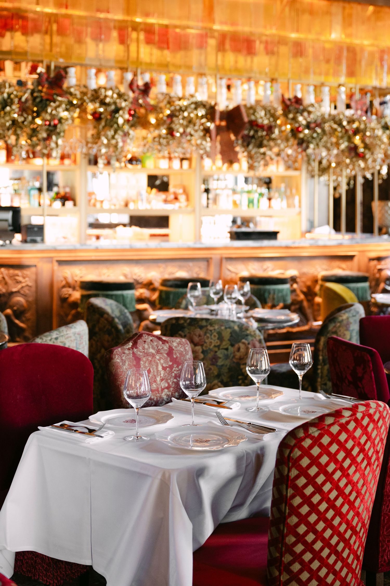 Upscale restaurant interior with red velvet chairs, white tablecloths, crystal glasses, and festive floral bar decor.