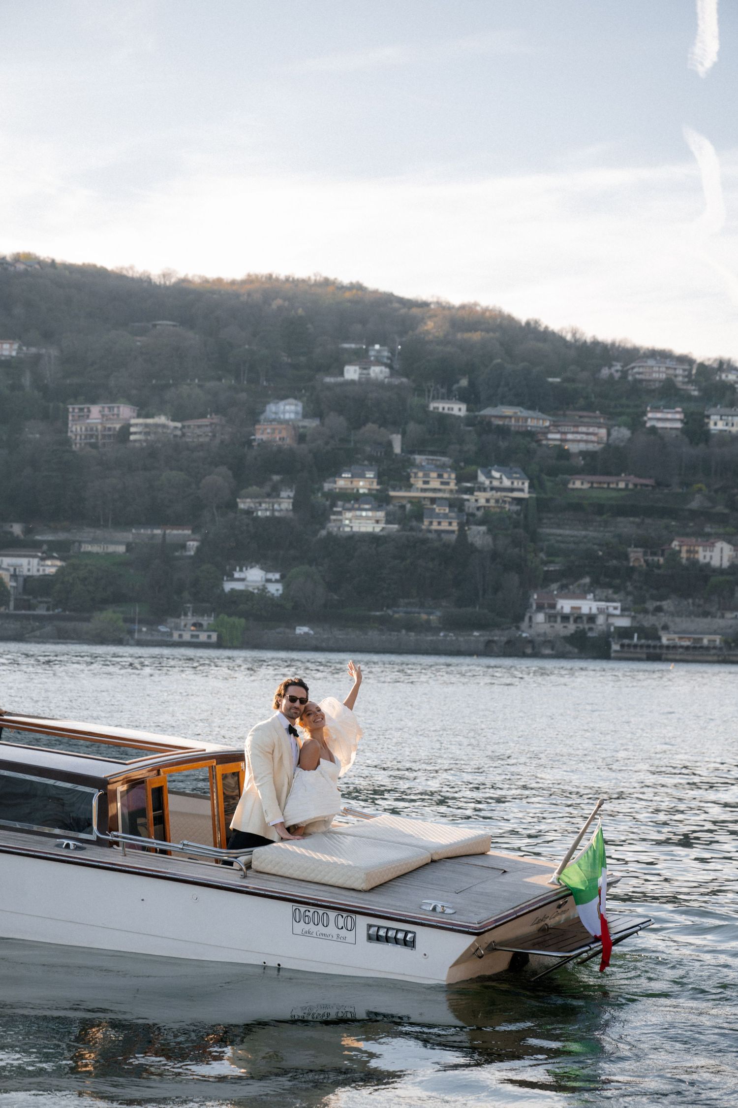 Bride and groom waving from classic Italian water taxi on Lake Como at golden hour.