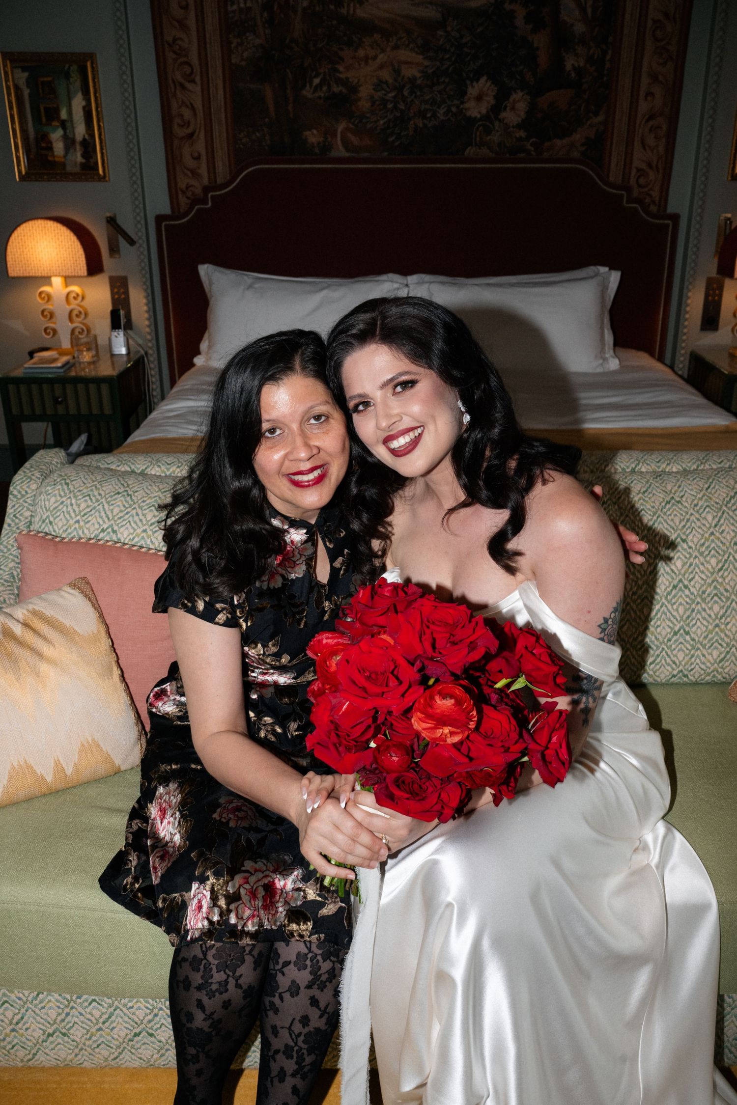 Bride in white satin gown and mother pose together smiling, bride holding lush red rose bouquet in hotel room.