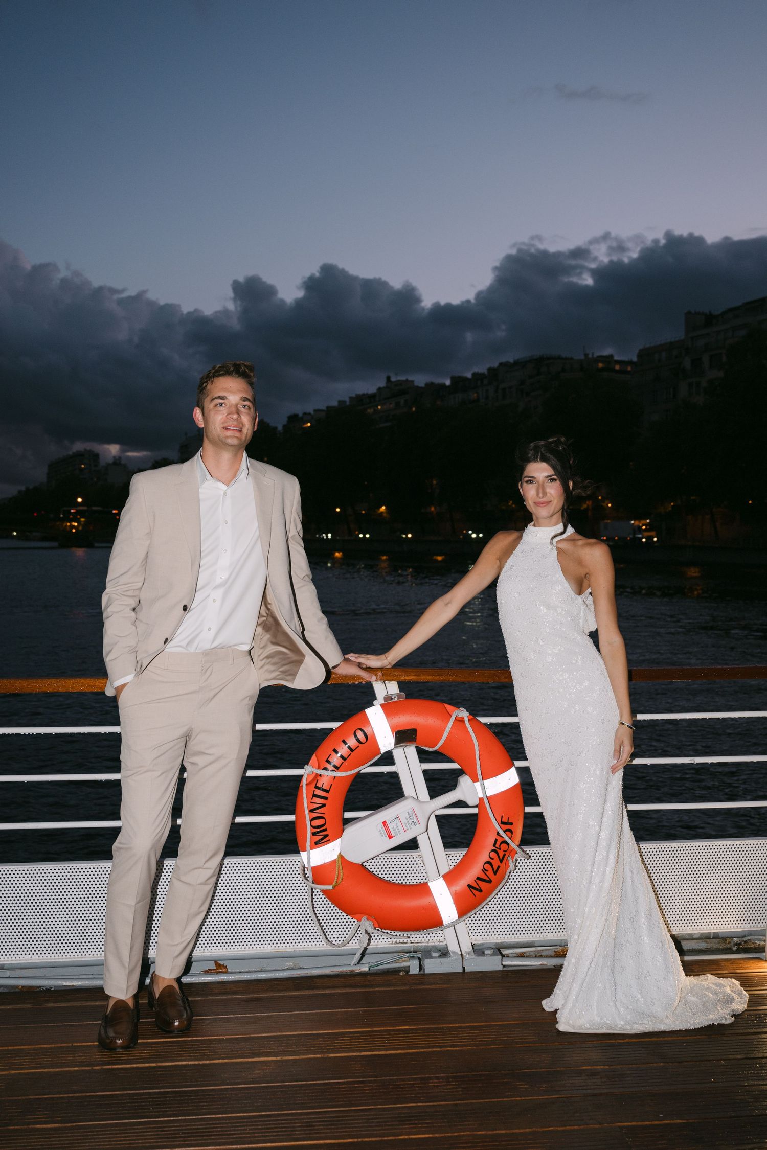 Bride and groom pose with a life preserver on a boat deck at dusk with coastal town lights in background.