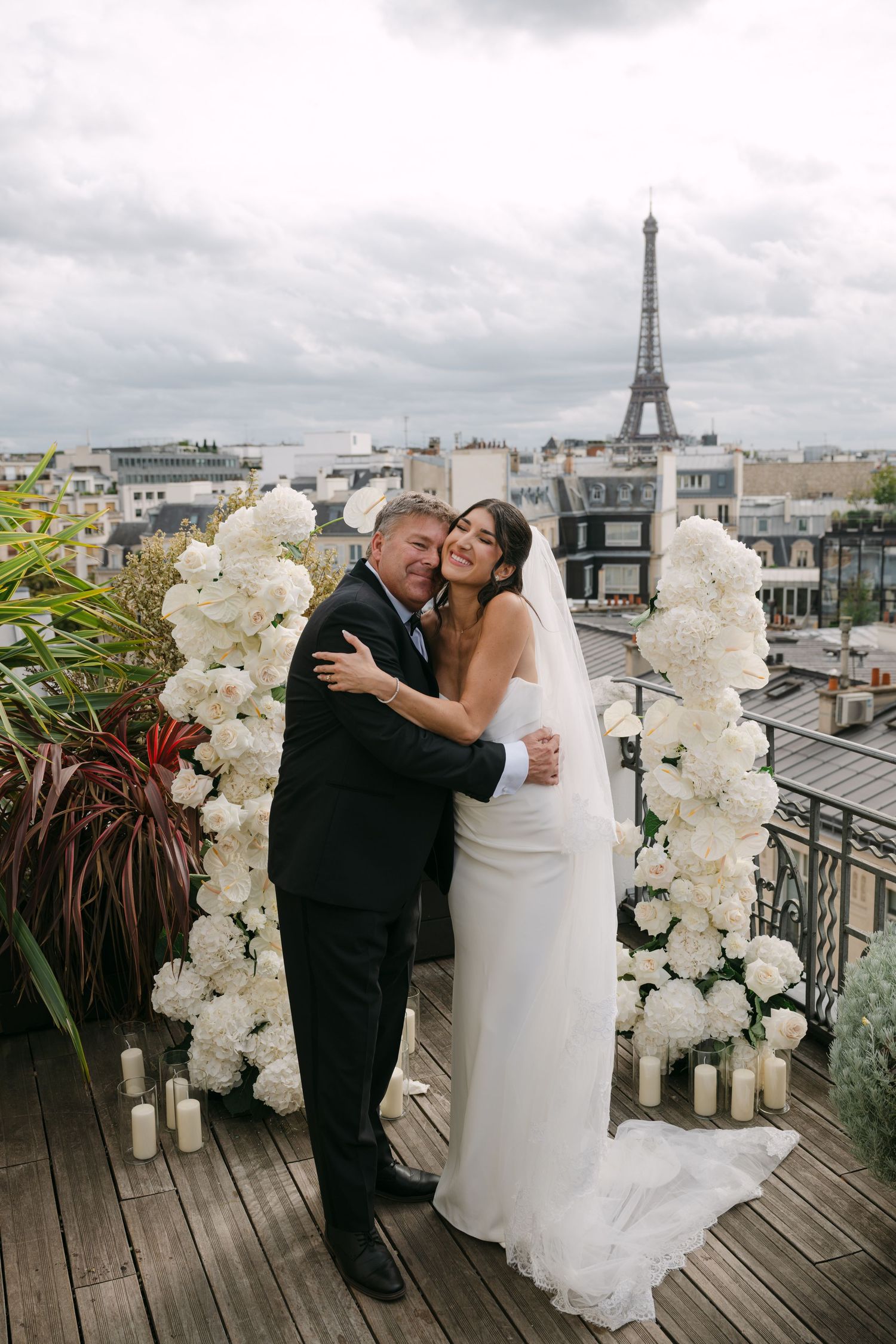 Bride and groom embracing at a rooftop wedding ceremony with white floral arch and Eiffel Tower backdrop.