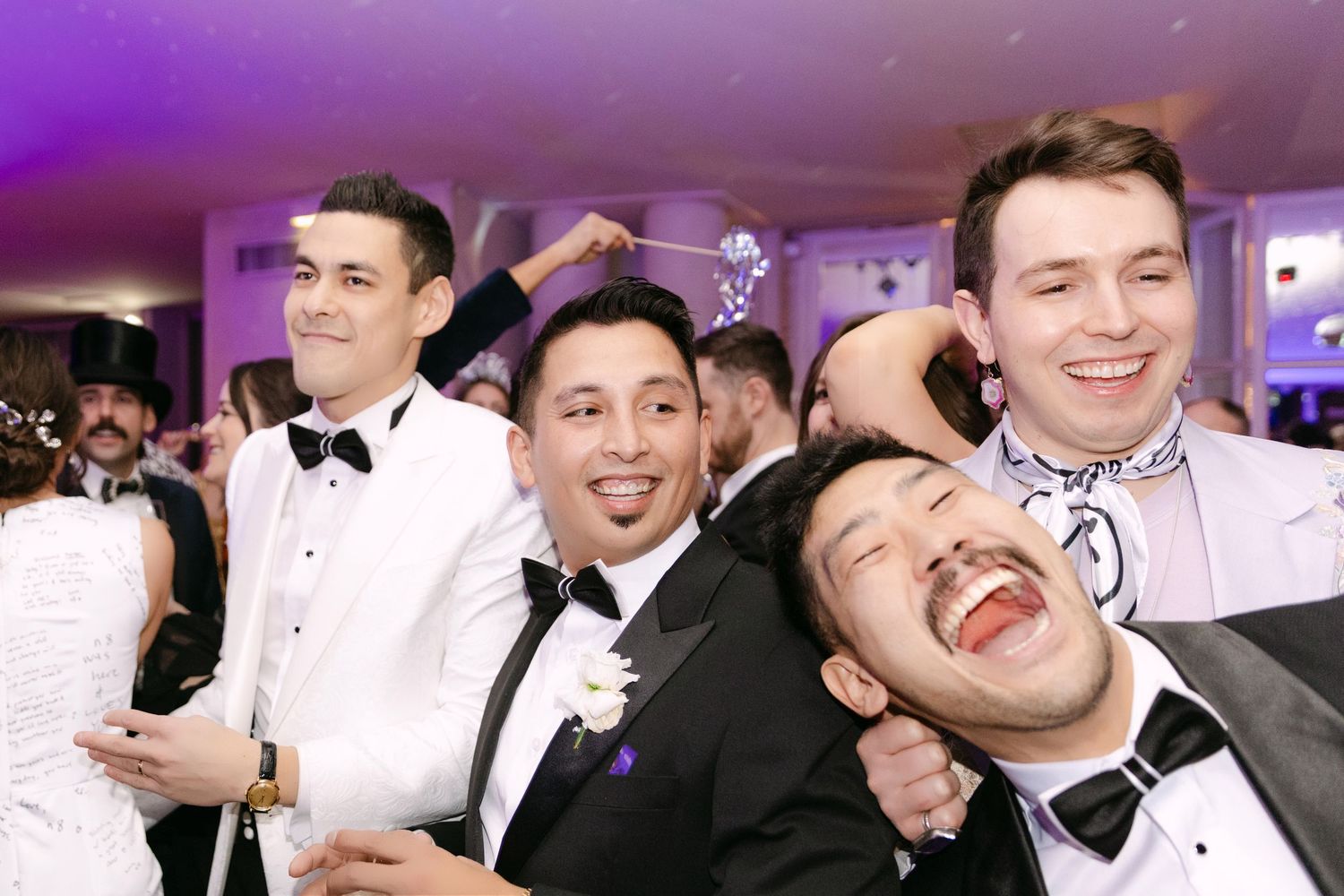 Four groosmen in tuxedos laugh and celebrate joyfully on the dance floor at a wedding reception.