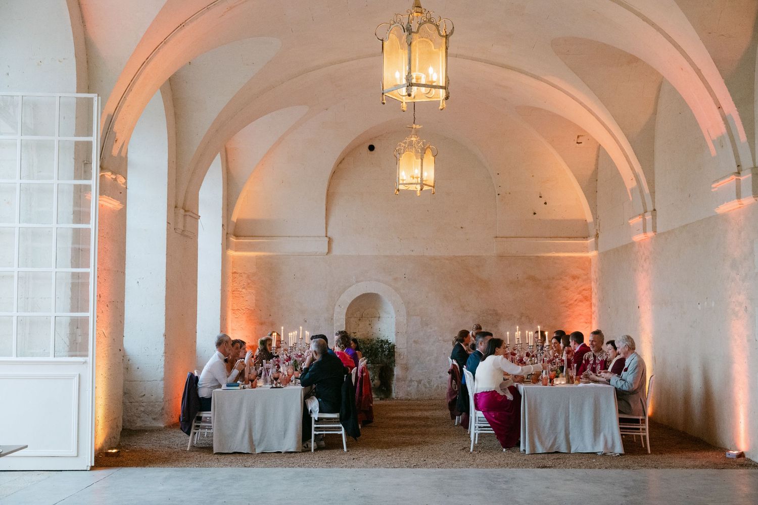 Wide shot of intimate wedding reception dinner in a warmly lit vaulted stone hall.