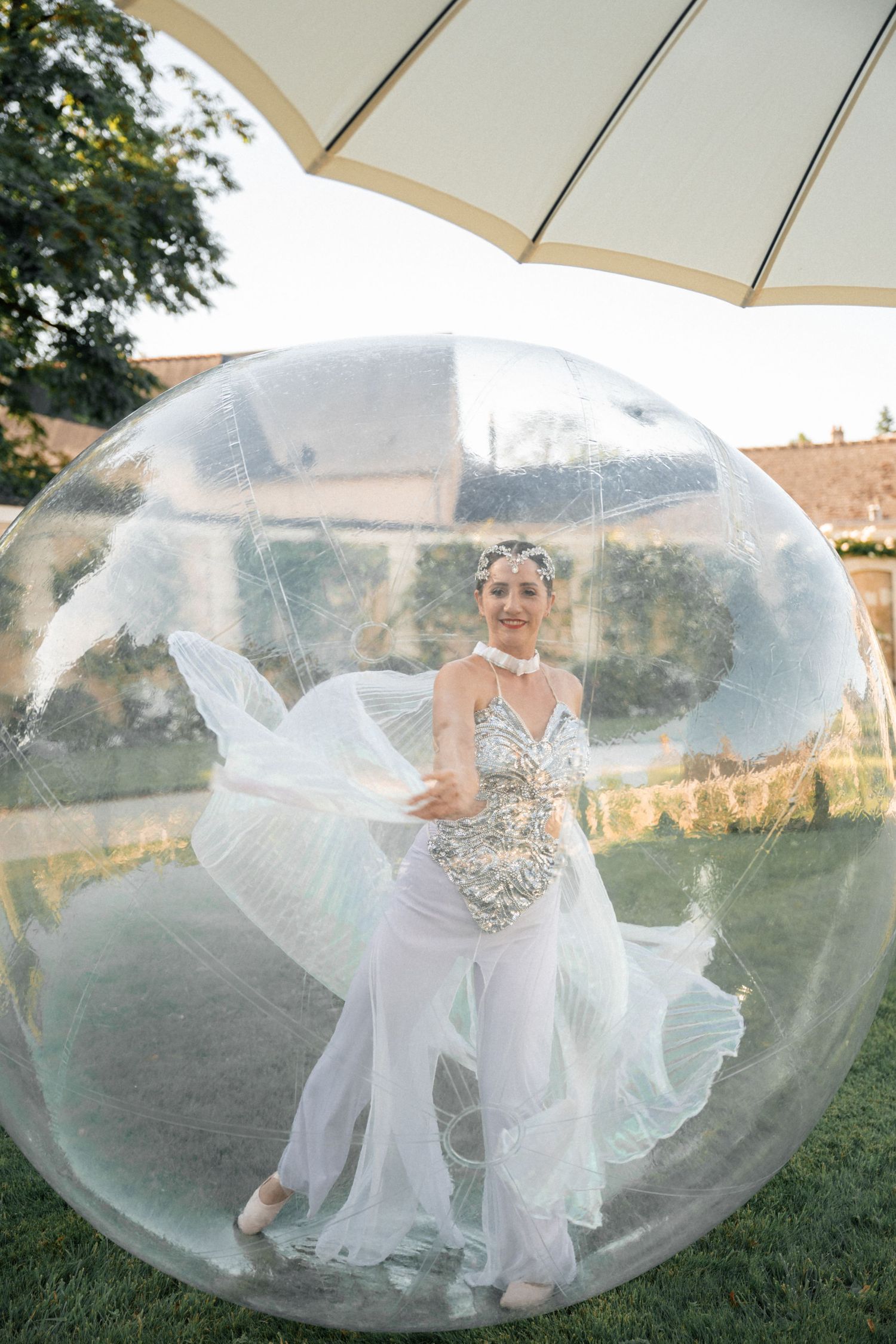 Smiling performer in a sparkling white costume dancing inside a large transparent inflatable bubble outdoors.