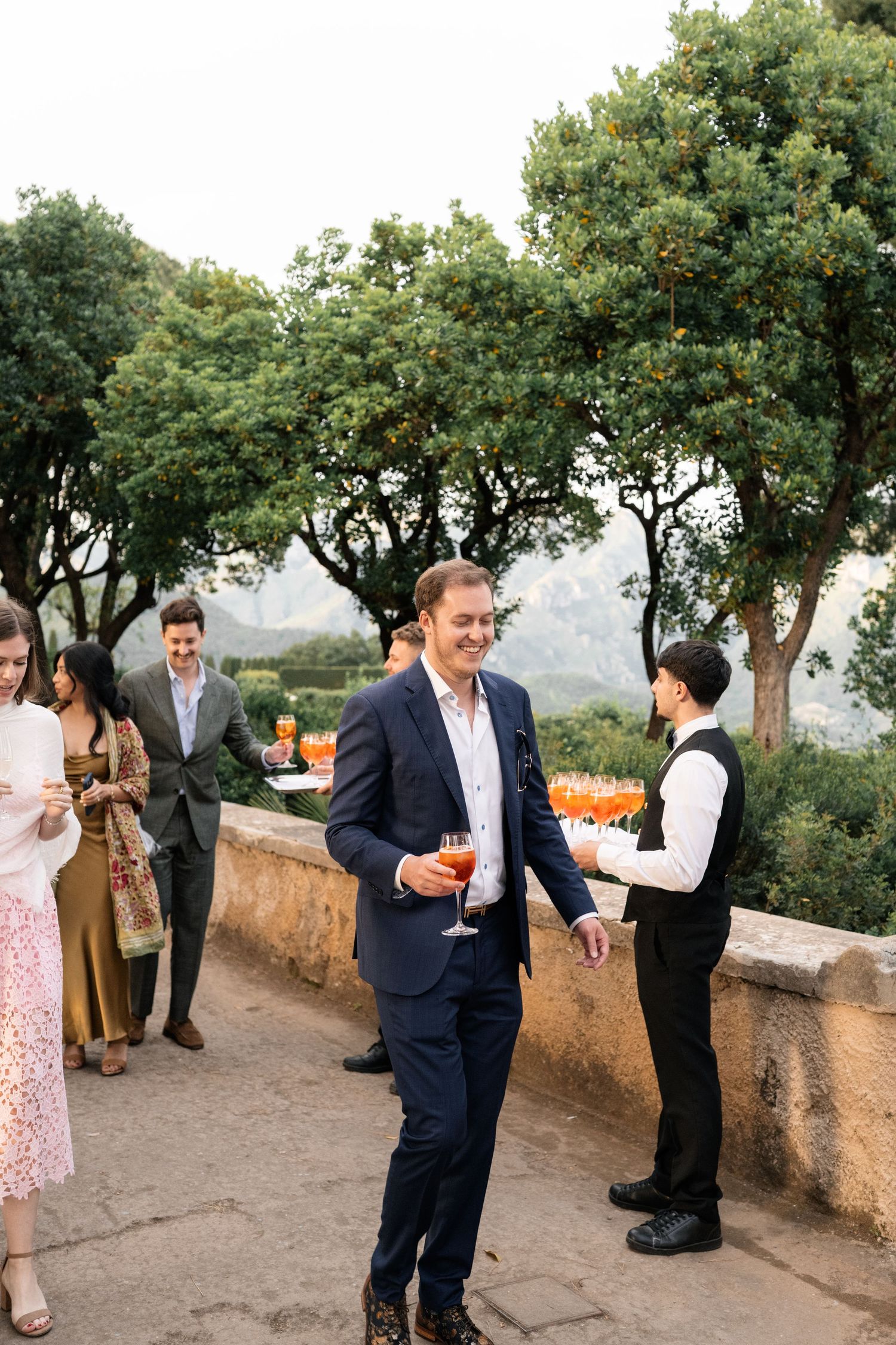 Guests mingle on a terrace during a cocktail hour, with a waiter serving Aperol Spritz drinks at an outdoor wedding reception.