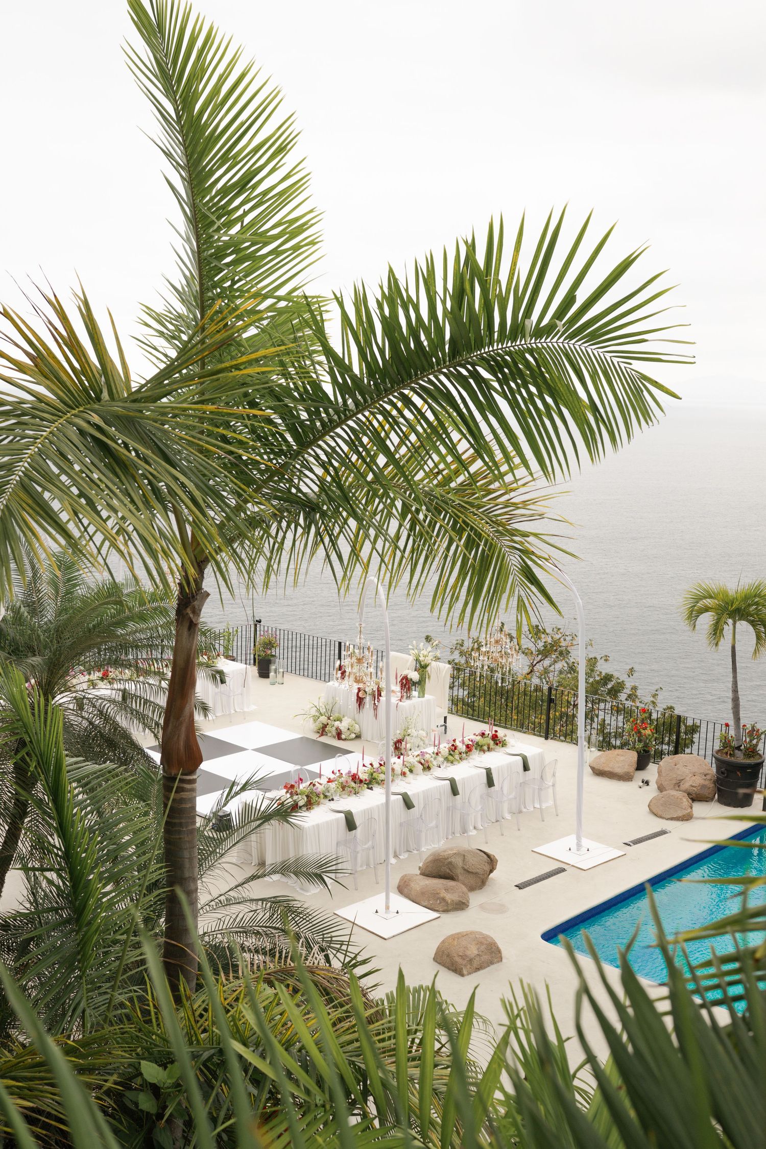 Elegant outdoor wedding reception setup with long white tables by a pool, overlooking the ocean, surrounded by lush palm trees.