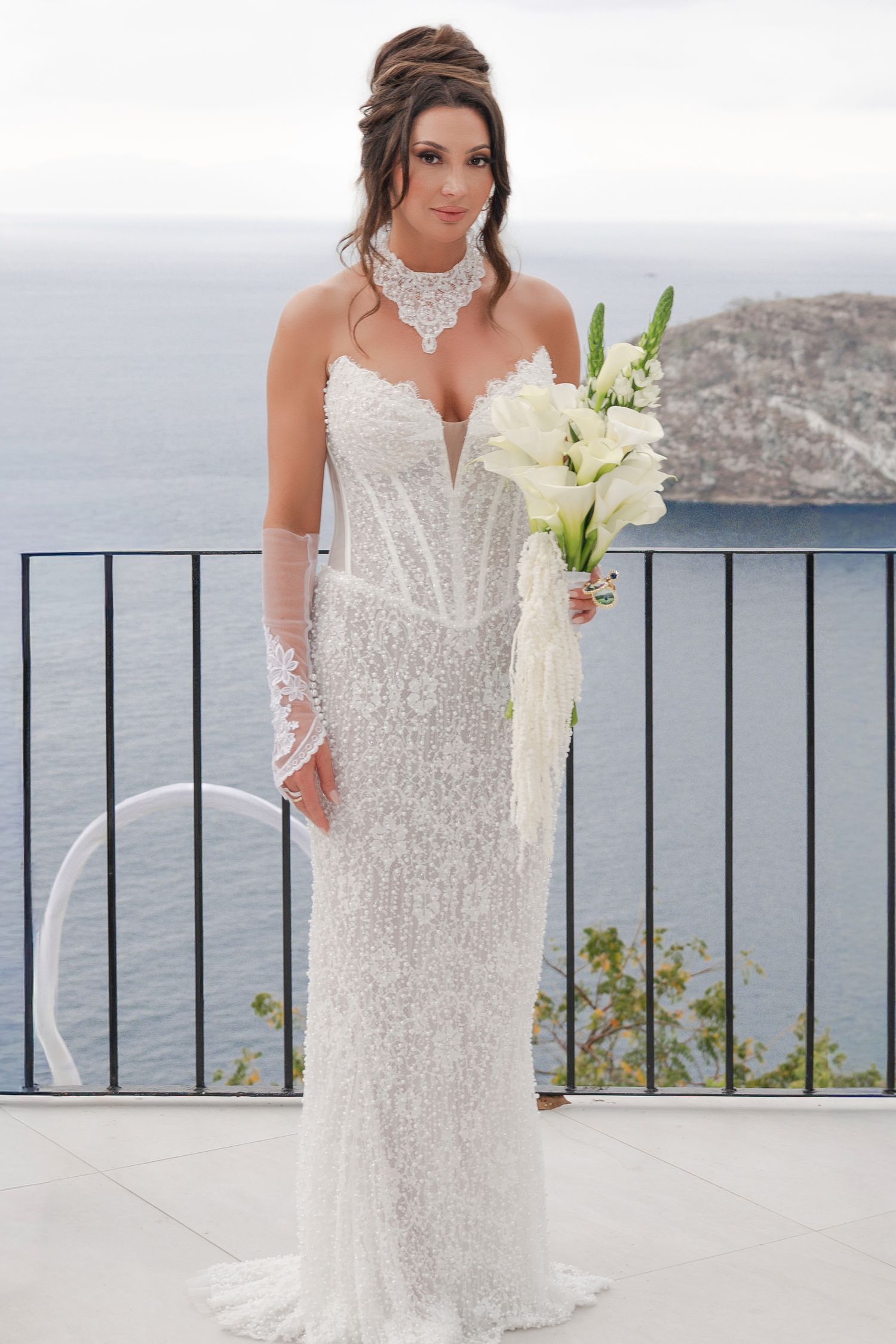 Bride in white lace gown holding bouquet of white flowers on balcony overlooking ocean.