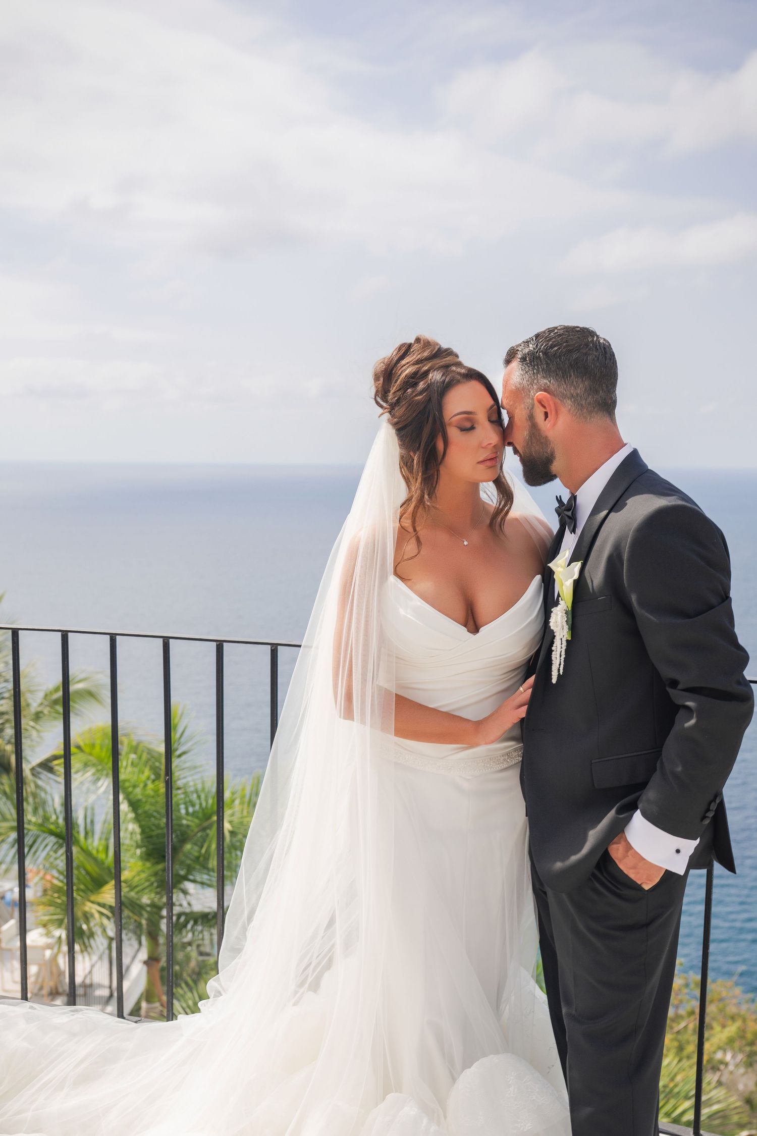 Newlyweds embracing tenderly on balcony overlooking tropical garden and blue ocean horizon.