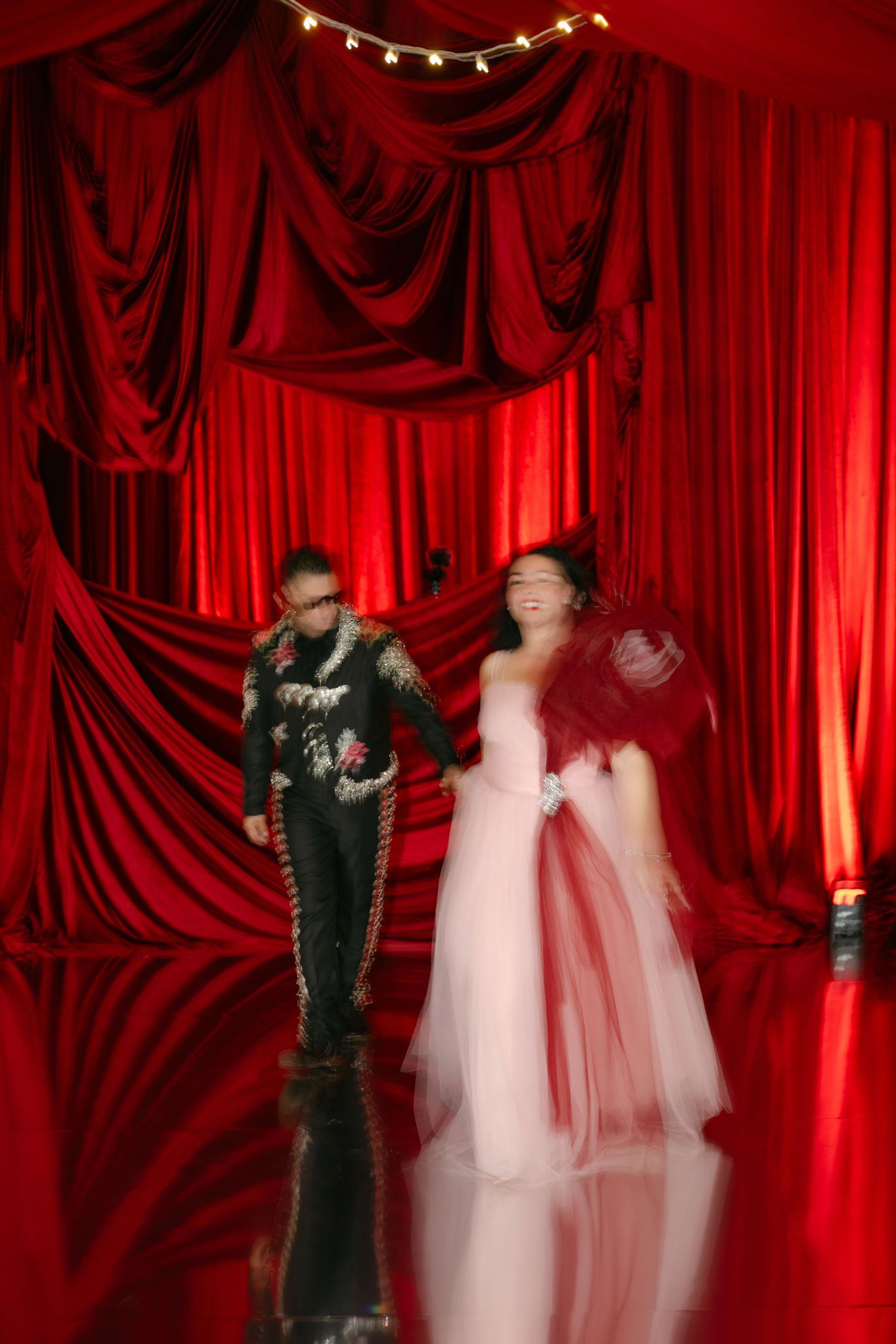 Couple in formal attire poses before dramatic red velvet curtains, woman in pink gown, man in dark suit.