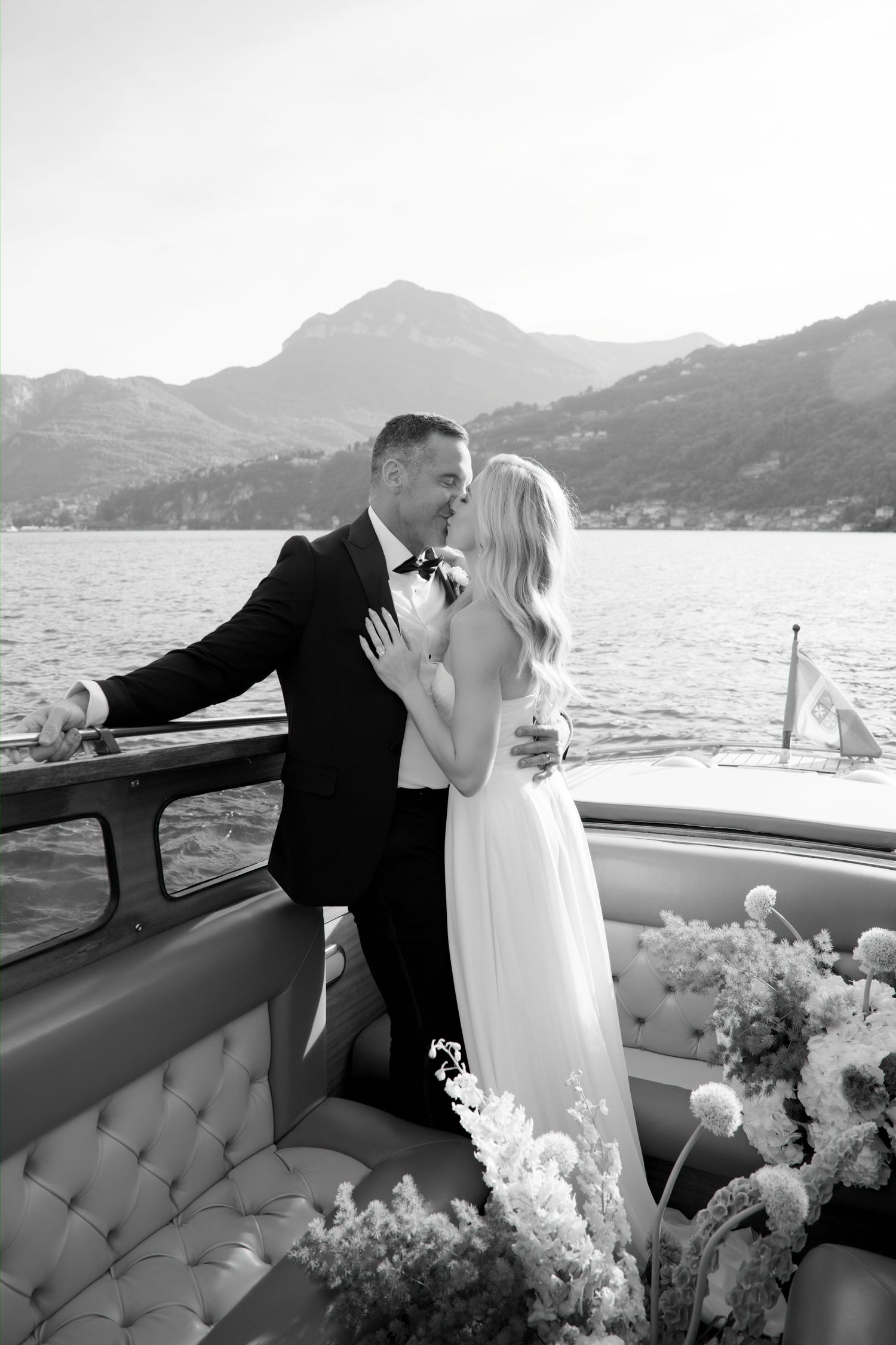 Black and white photo of a bride and groom kissing on a luxury boat with mountain lake views.