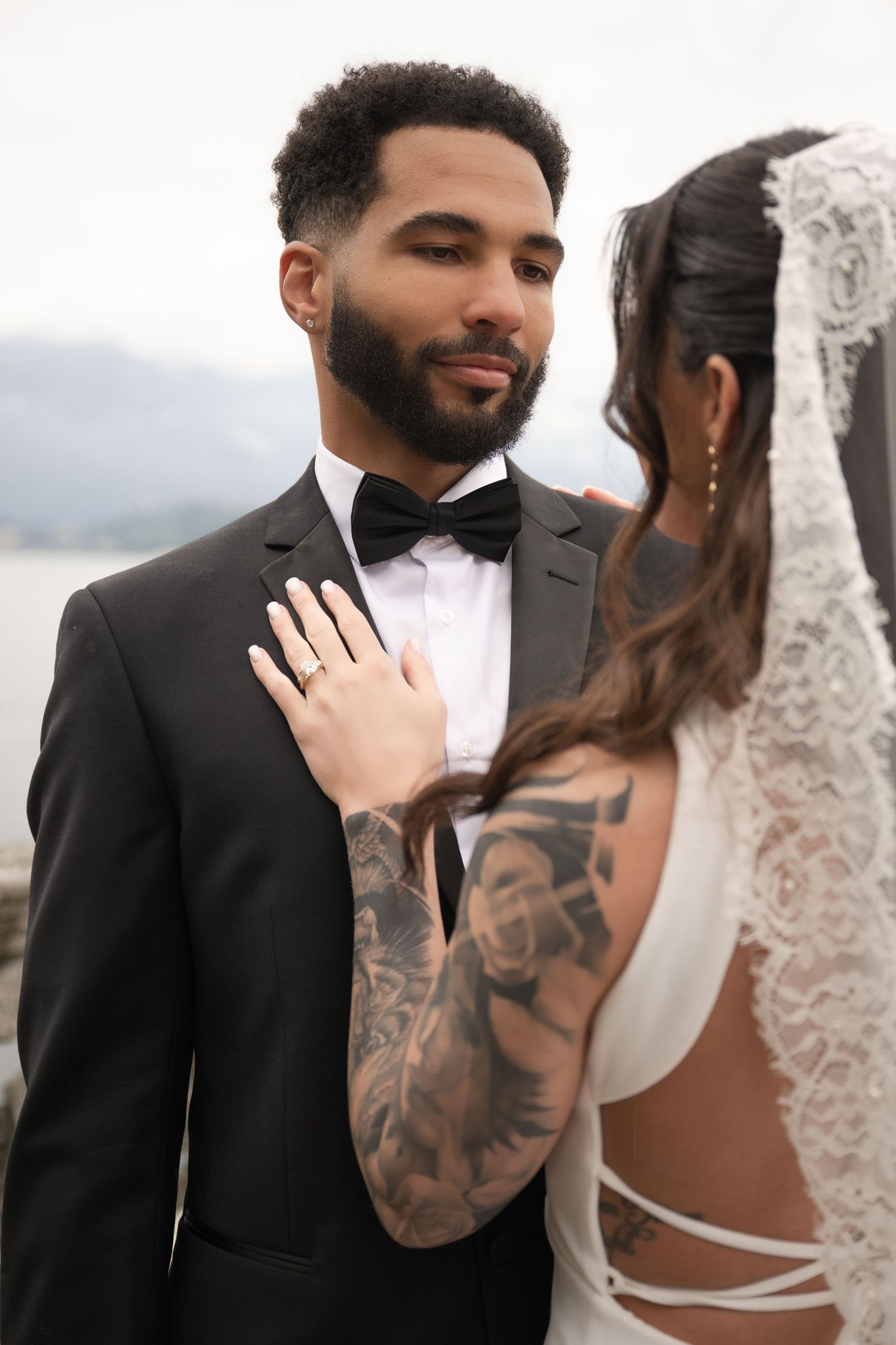 Bride with tattoos and wedding ring places hand on groom in black tuxedo at outdoor waterfront ceremony.