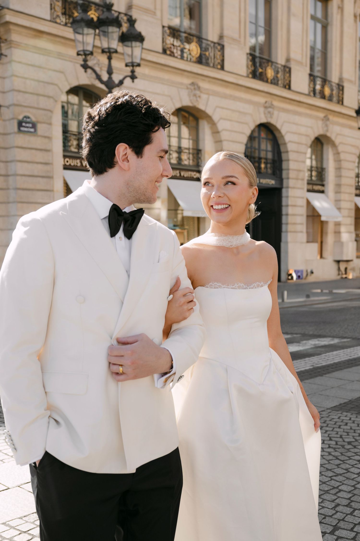 Smiling bride in strapless white gown walks arm-in-arm with groom in white tuxedo near Louis Vuitton in Paris.