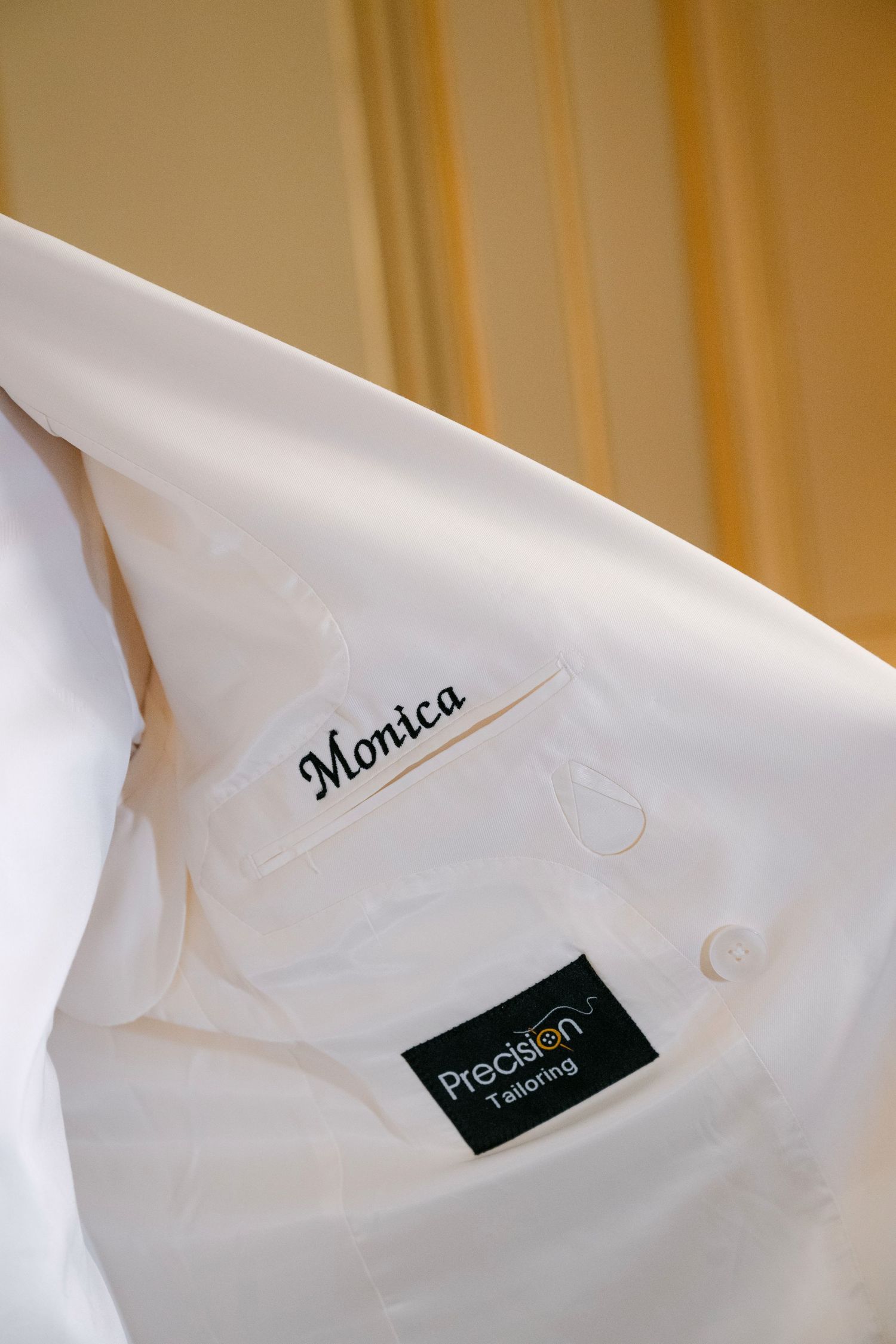 Custom white wedding jacket with embroidered name Monica by Precision Tailoring hangs on display.