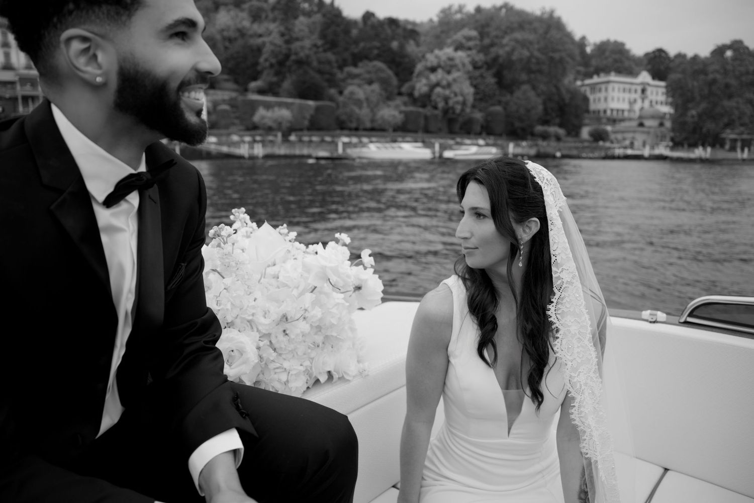 Black and white photo of a bride and groom on a boat, bride holding white flowers, scenic waterfront in background.