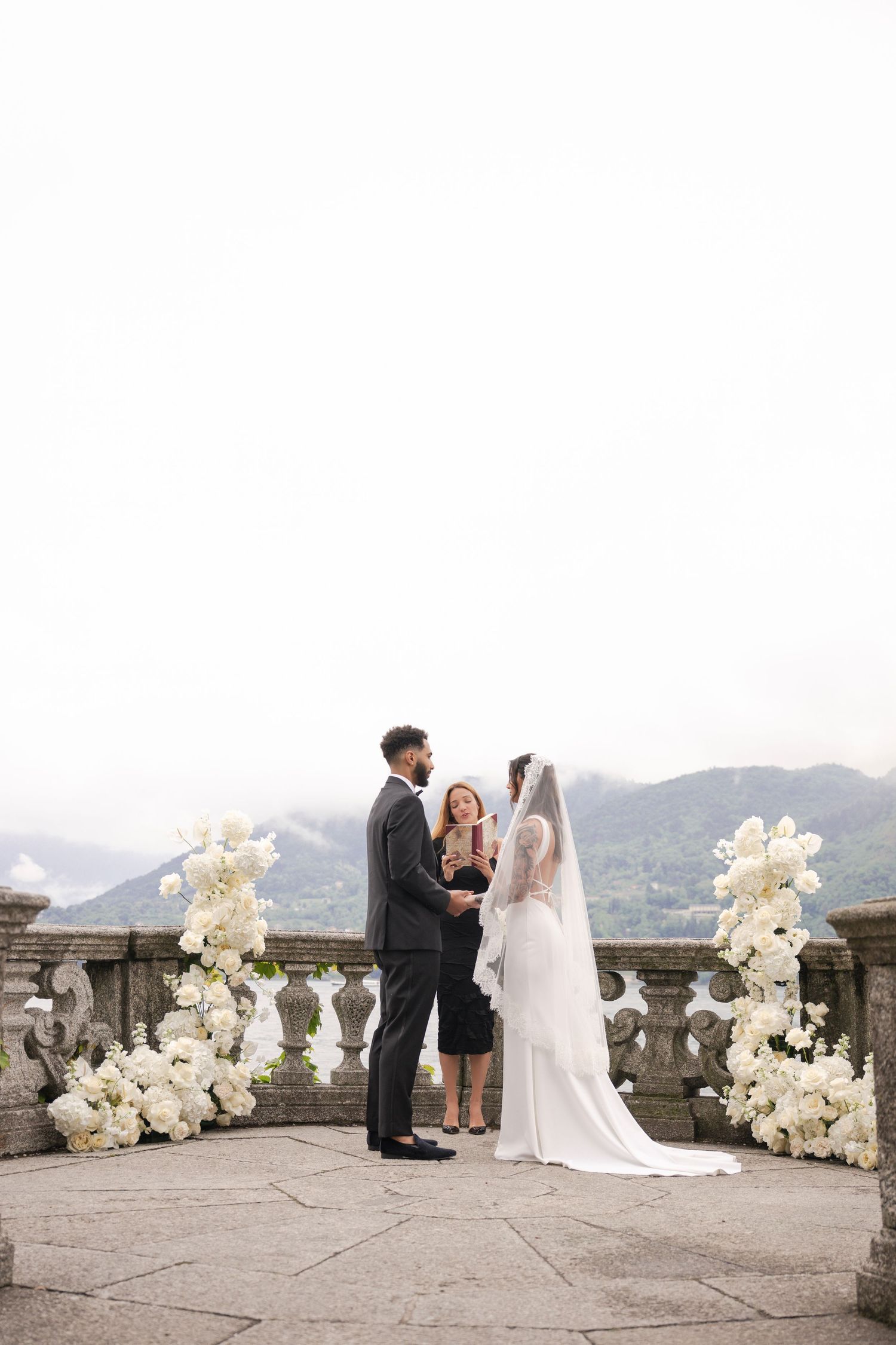 Couple exchanging vows on an elegant stone terrace adorned with white floral arrangements and mountain views.
