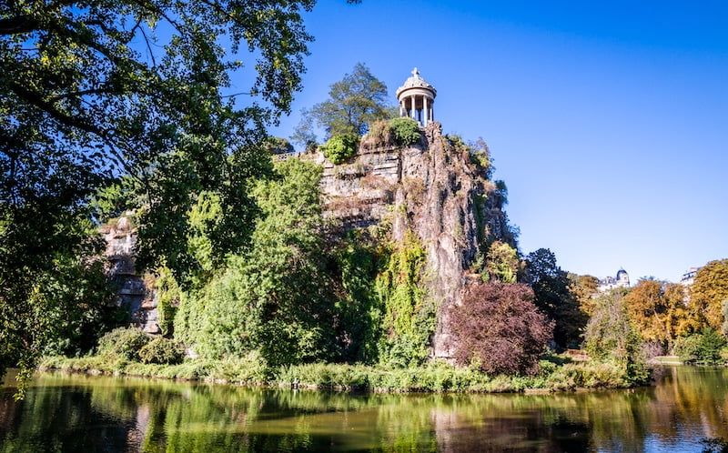 Temple-like structure perched on a rocky cliff overlooking a serene lake surrounded by lush trees.