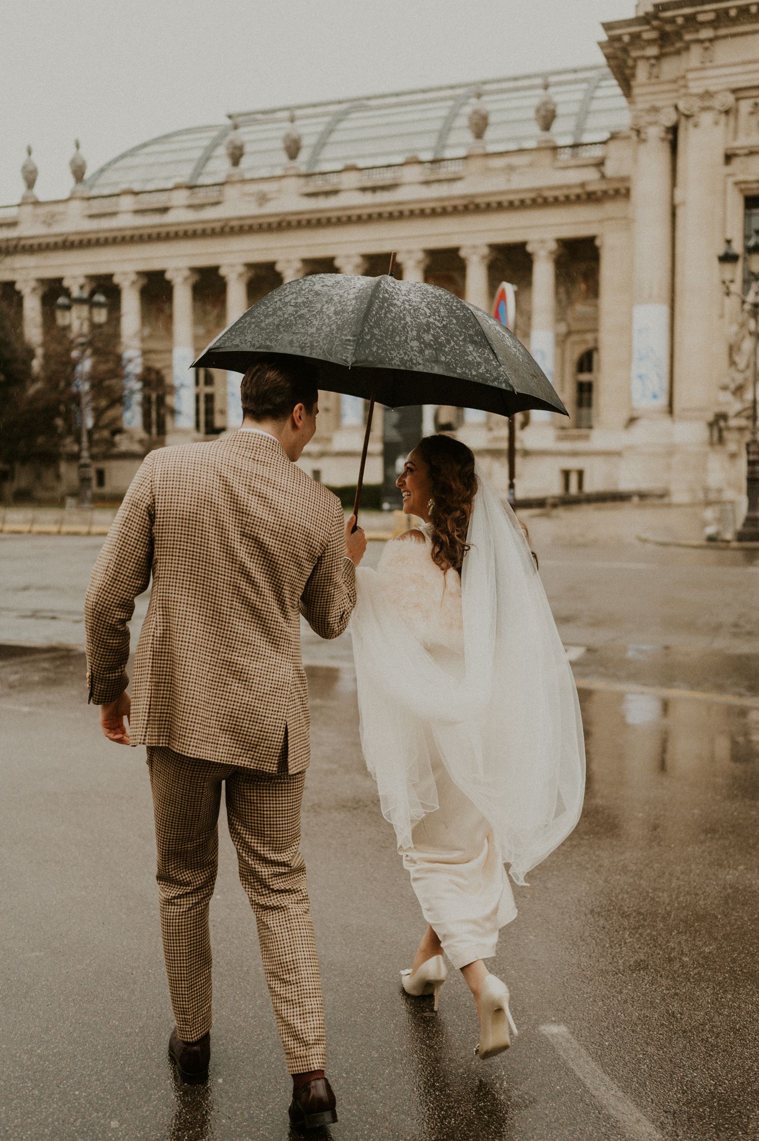 Sepia-toned photograph of couple walking away sharing umbrella near classical architecture.