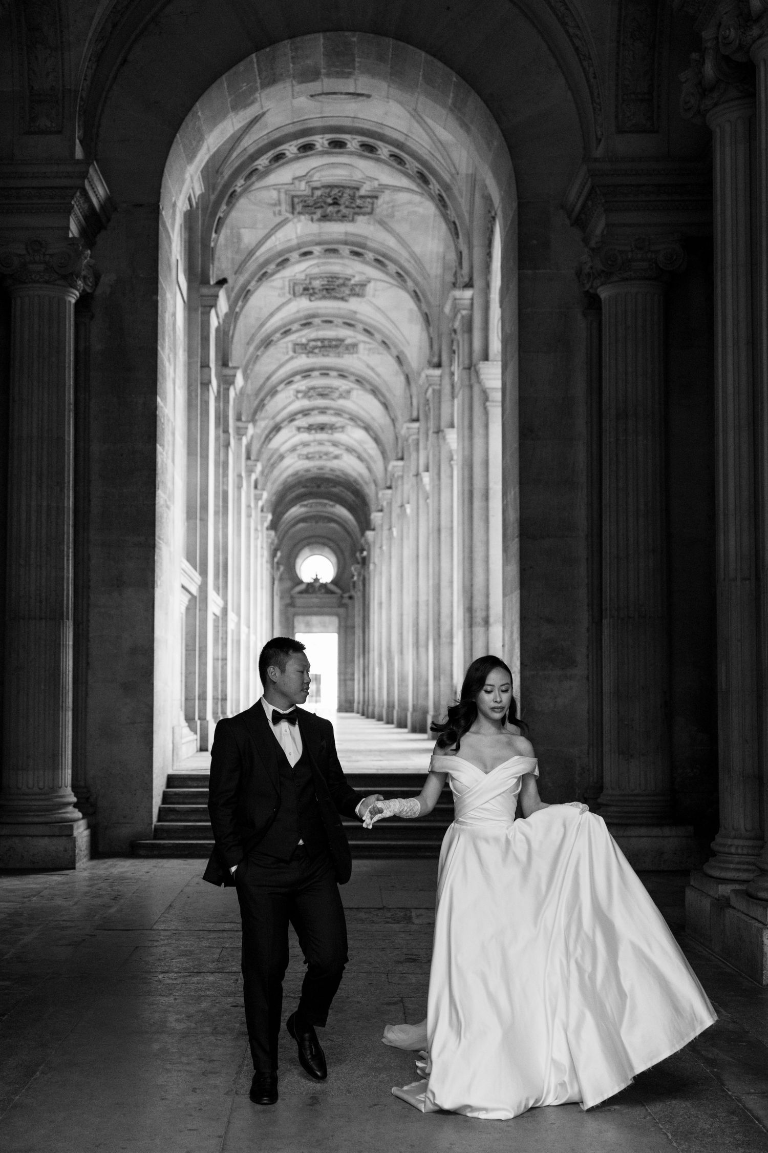 Wedding couple walks hand in hand through grand arched colonnade in black and white portrait.