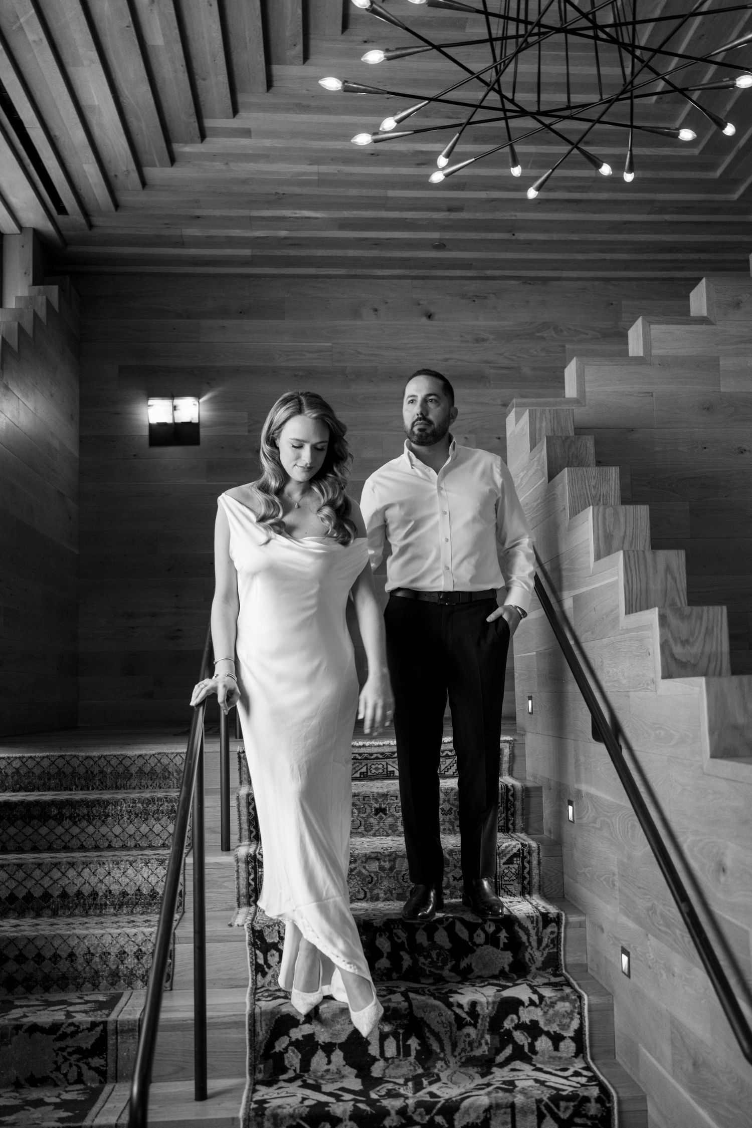 Elegant couple descending ornate staircase in black and white, woman in white gown, man in dress shirt.