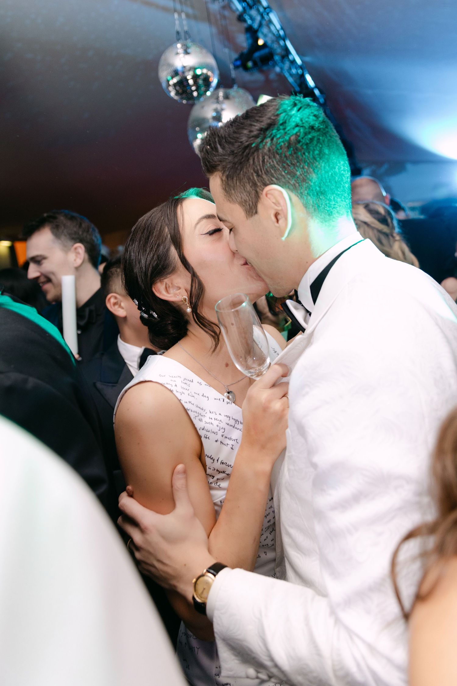 Bride and groom kiss on dance floor under disco balls while groom holds champagne glass.