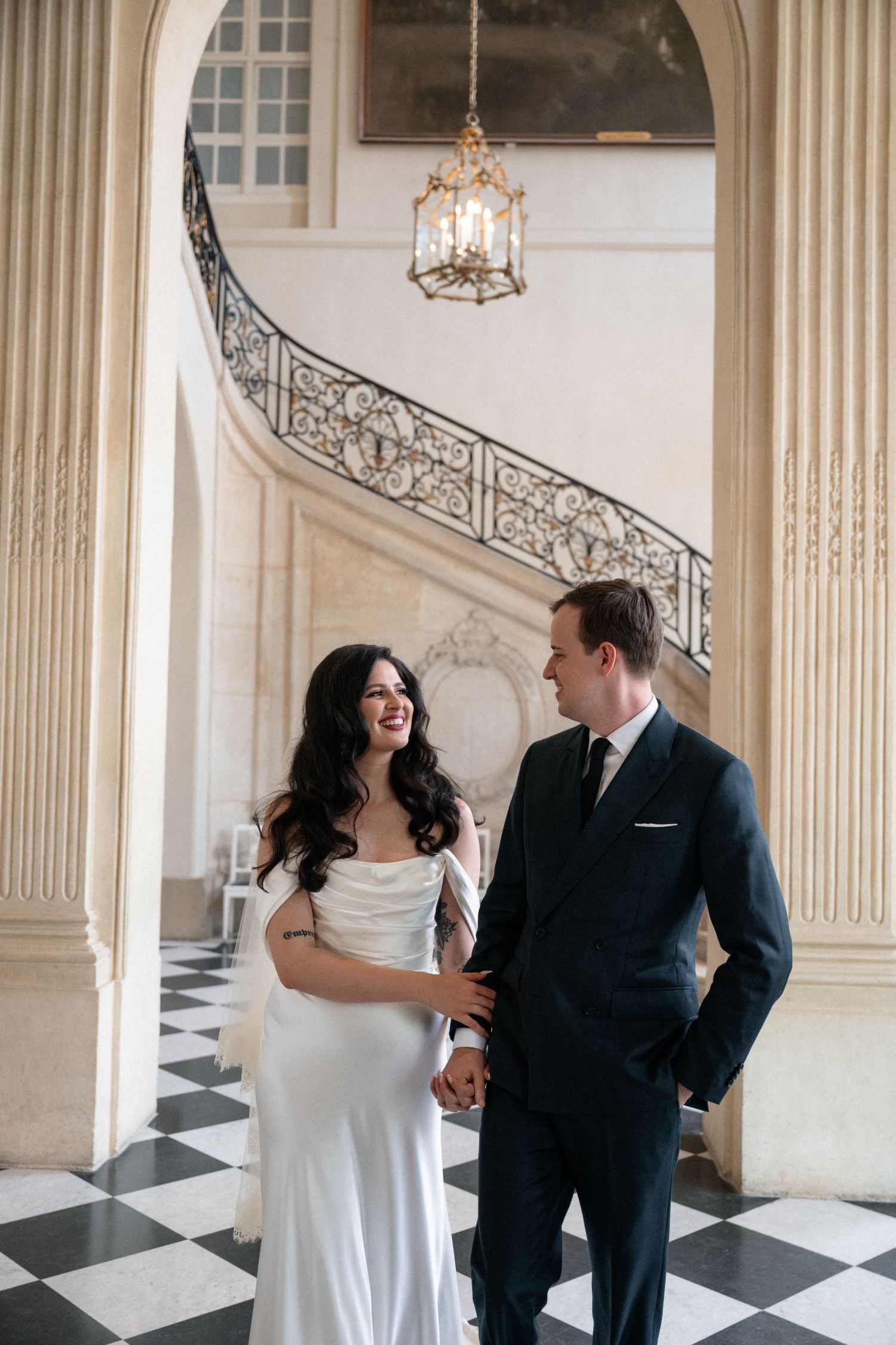 Bride and groom laughing together in elegant Parisian chateau with checkered floors and ornate spiral staircase.
