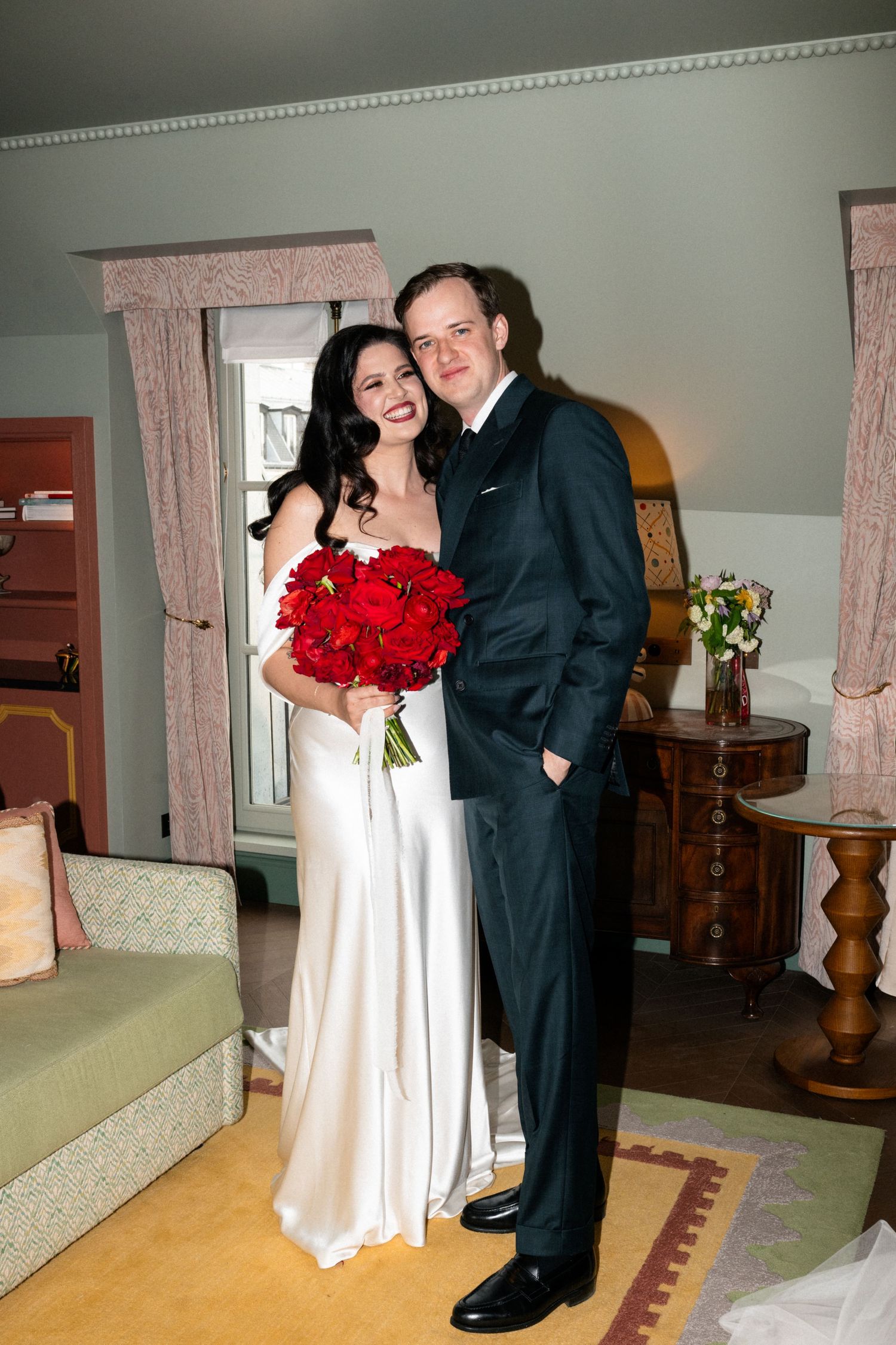 Bride and groom pose together smiling in elegant hotel suite, bride holding vibrant red rose bouquet.