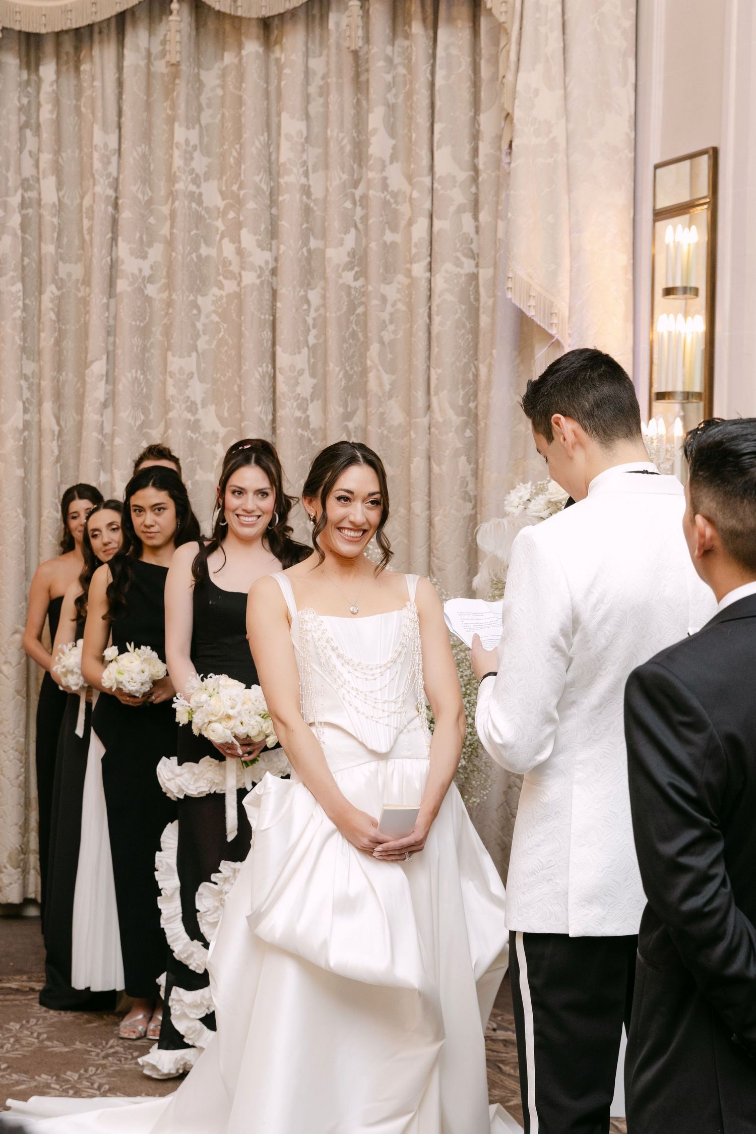 Smiling bride in pearl-embellished gown listening to groom read vows, bridesmaids in black dresses behind her.